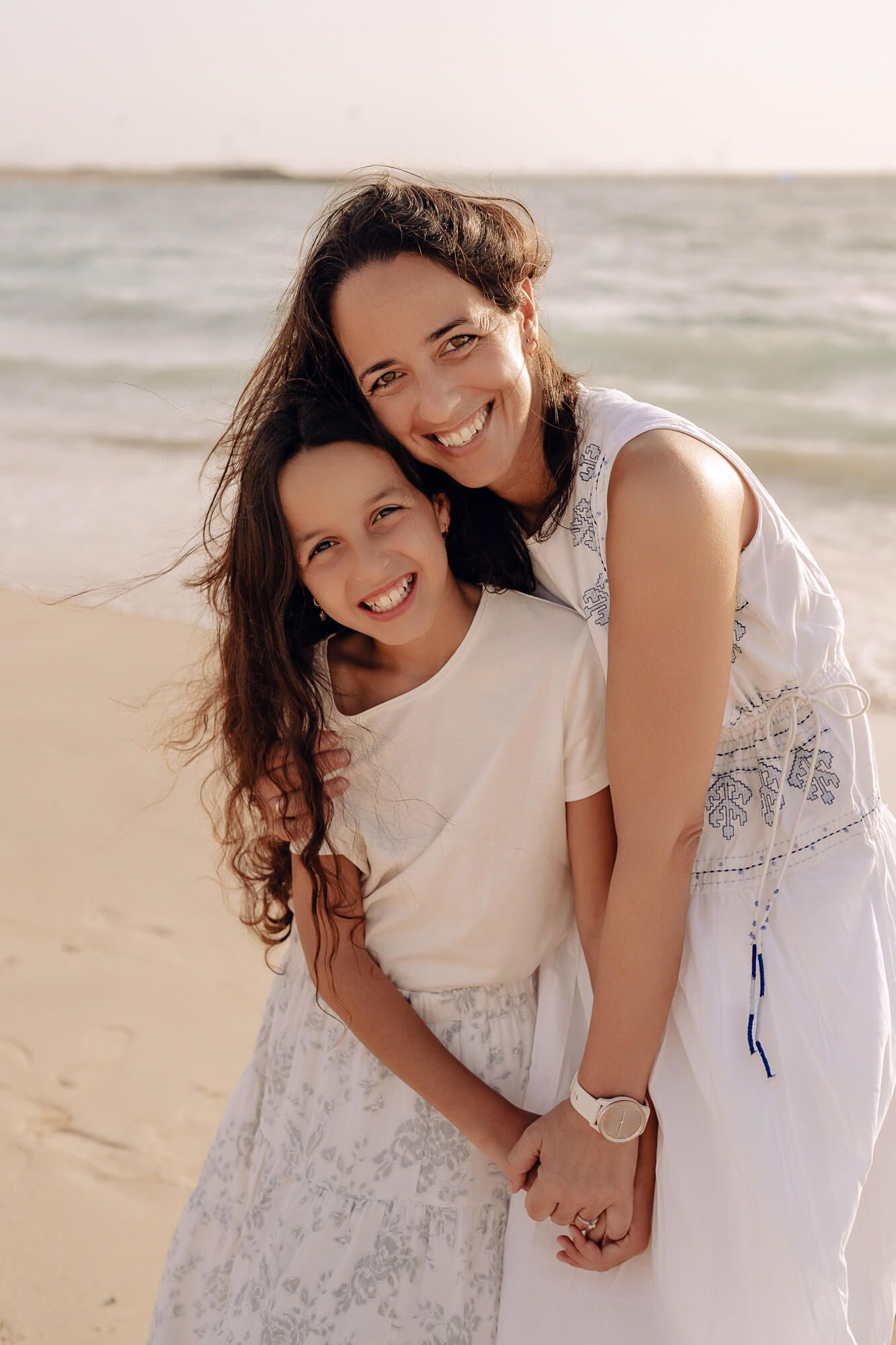 Two people in white dresses share a joyful embrace on a sunny beach with ocean waves in the background.