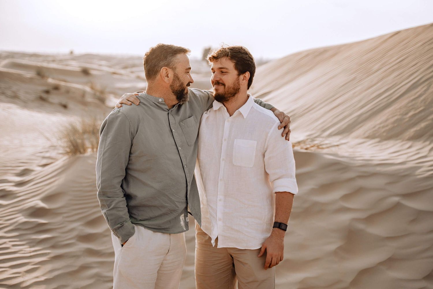 Two friends share a candid moment together against a backdrop of rolling desert dunes.