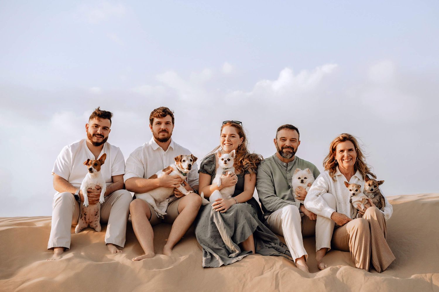 Casual desert photoshoot of group sitting on sand dunes with small dogs in neutral colored outfits.