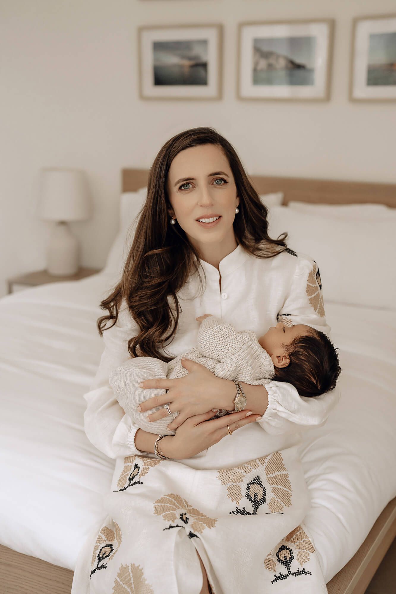 Mother in white dress holding sleeping baby during intimate lifestyle photoshoot on bed.