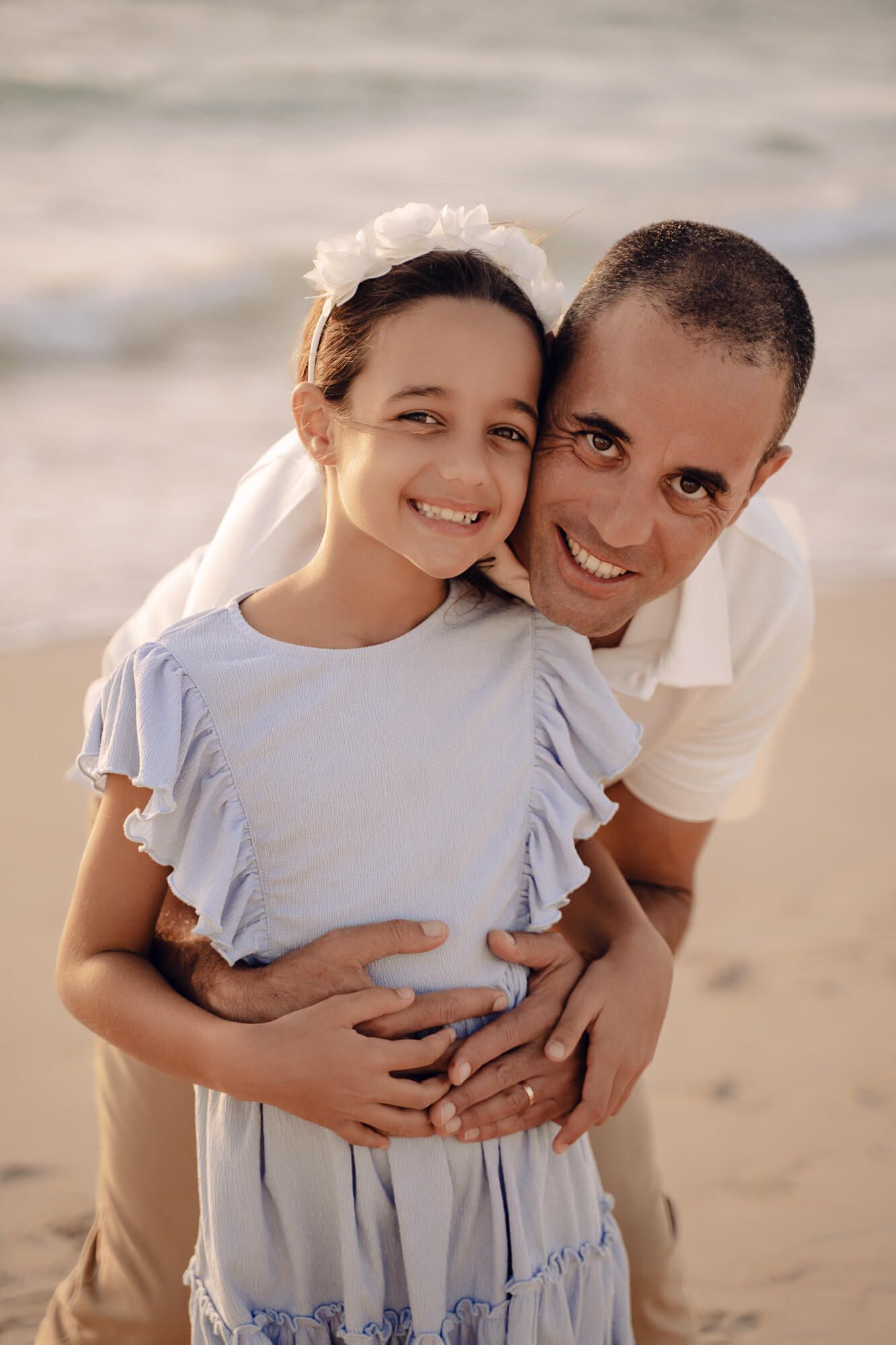 Tender family moment captured on beach with matching white and blue outfits at golden hour.