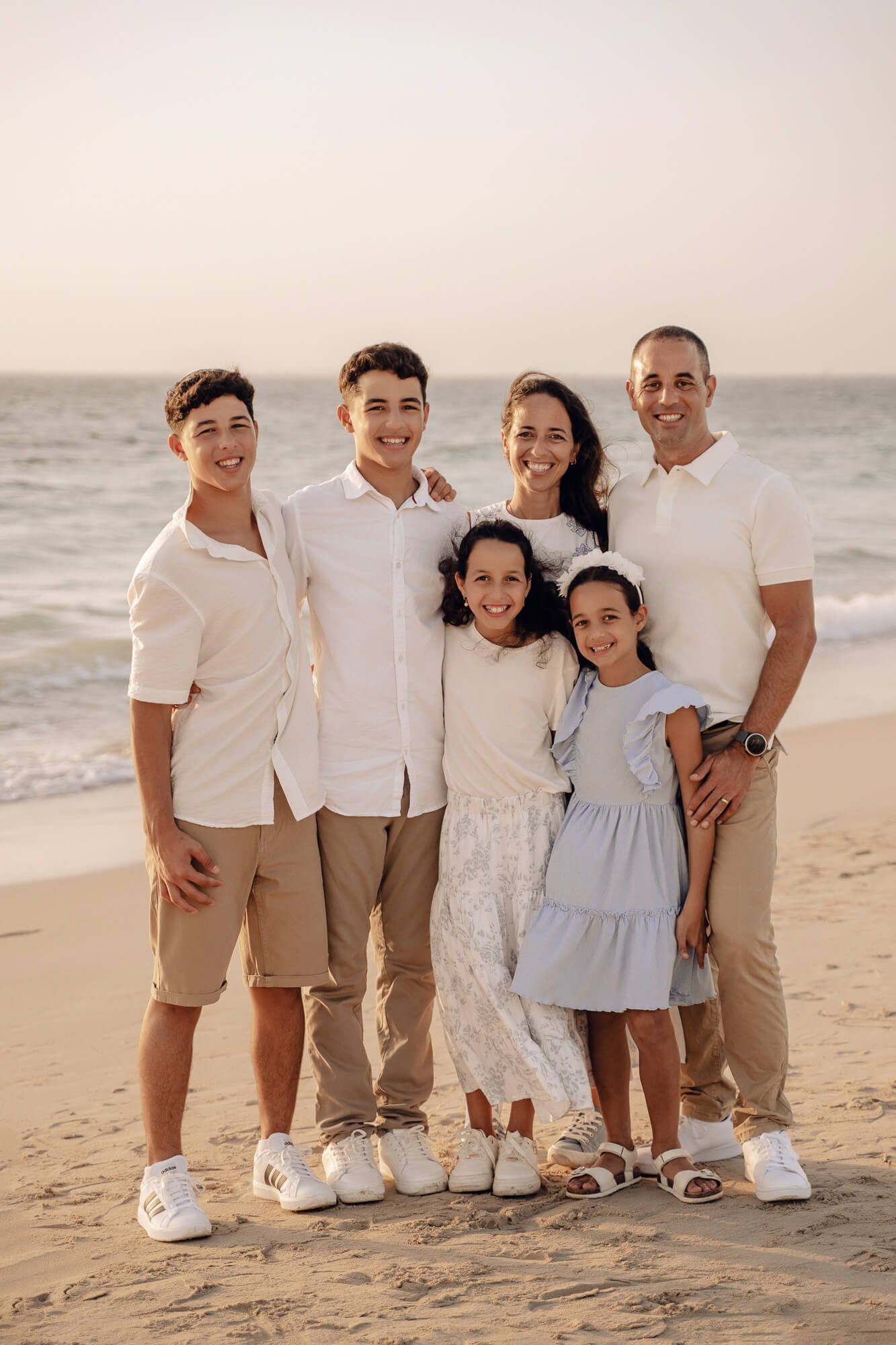 Family of six in matching white and beige outfits pose together on beach at sunset.
