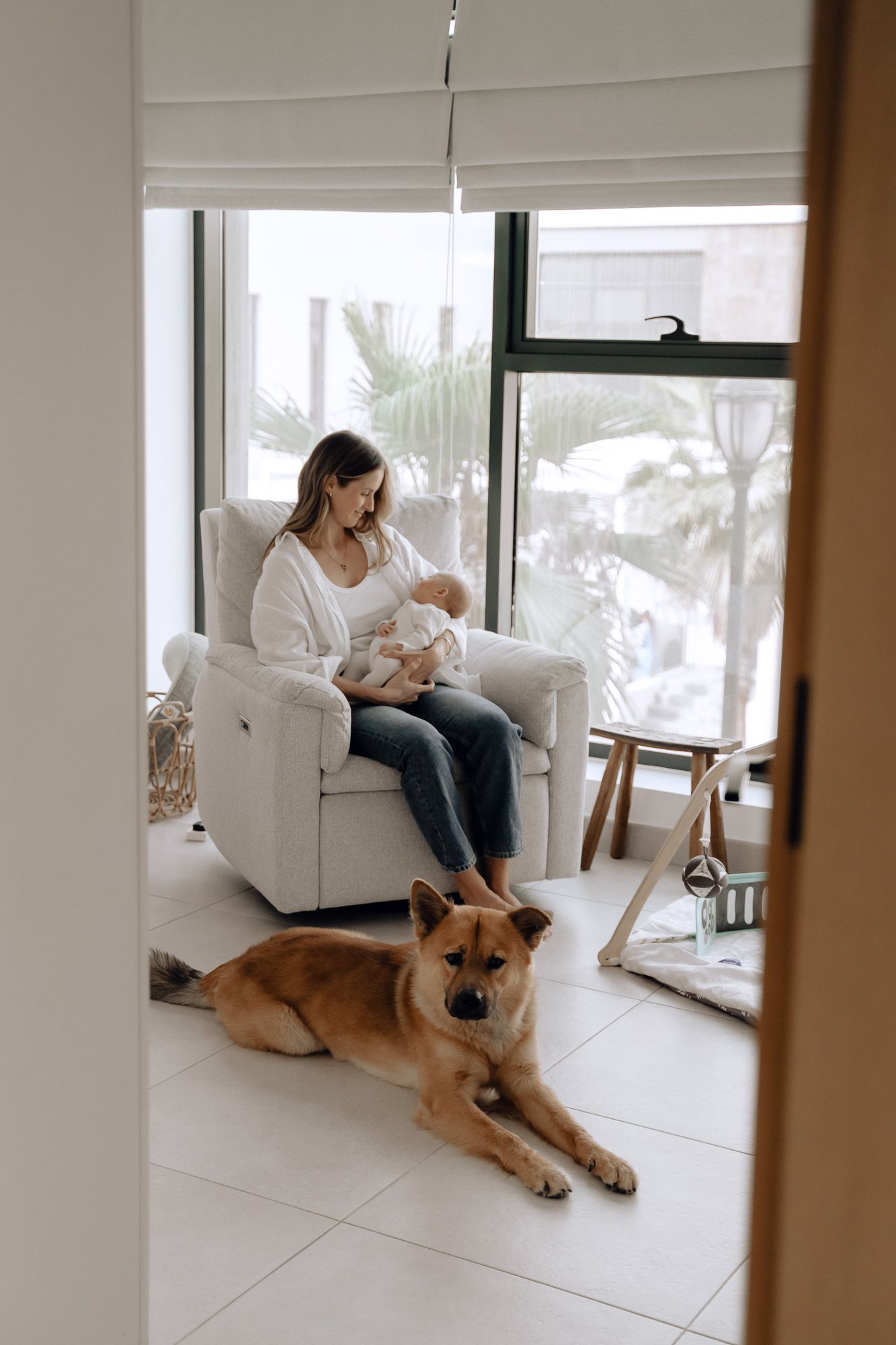 Woman sitting in white armchair with newborn while dog rests on floor by window.