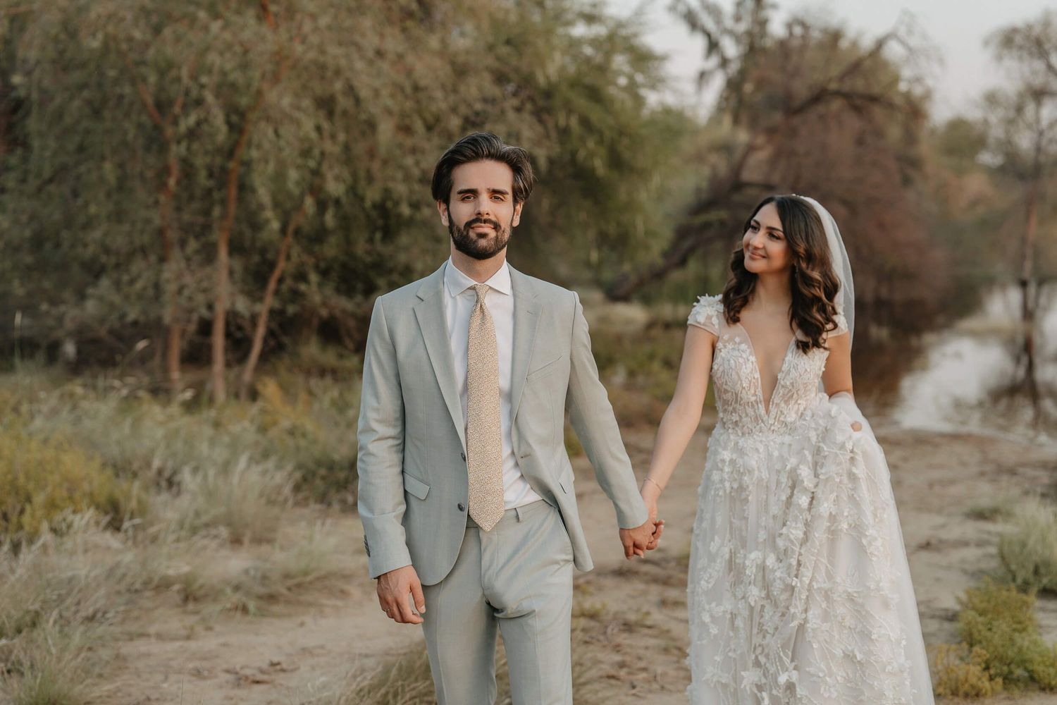 A couple in wedding attire walks through a natural outdoor setting during golden hour.