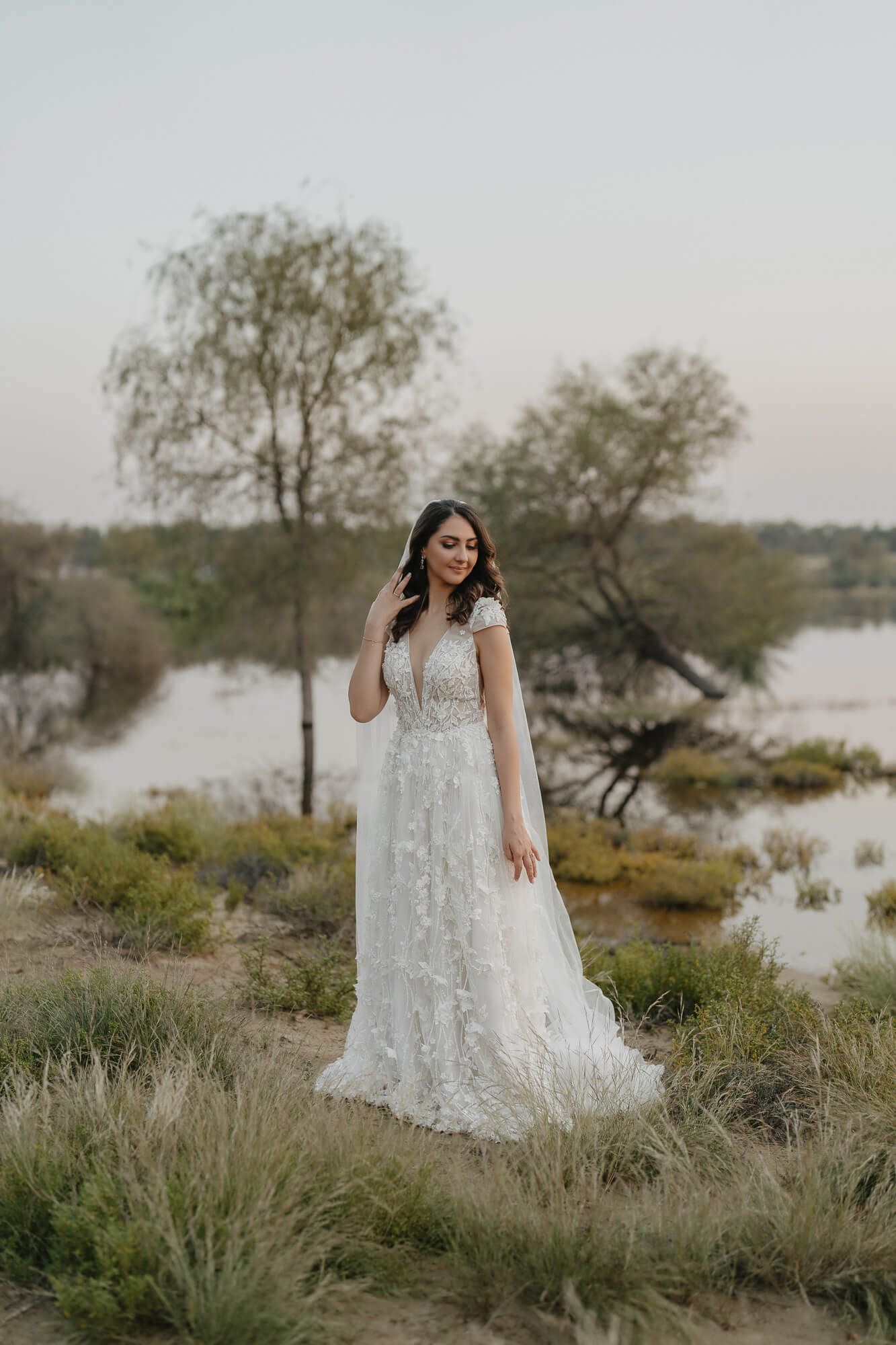 A bride in a lace wedding dress poses gracefully against a scenic desert background near a lake at sunset.