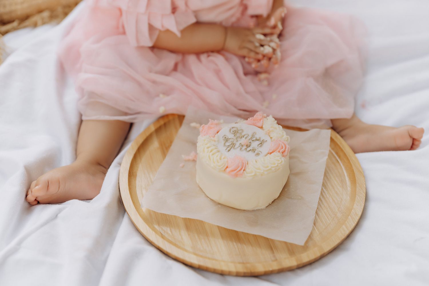 A small white birthday cake with pink decorations sits on a wooden plate beside pink tulle fabric on white bedding.