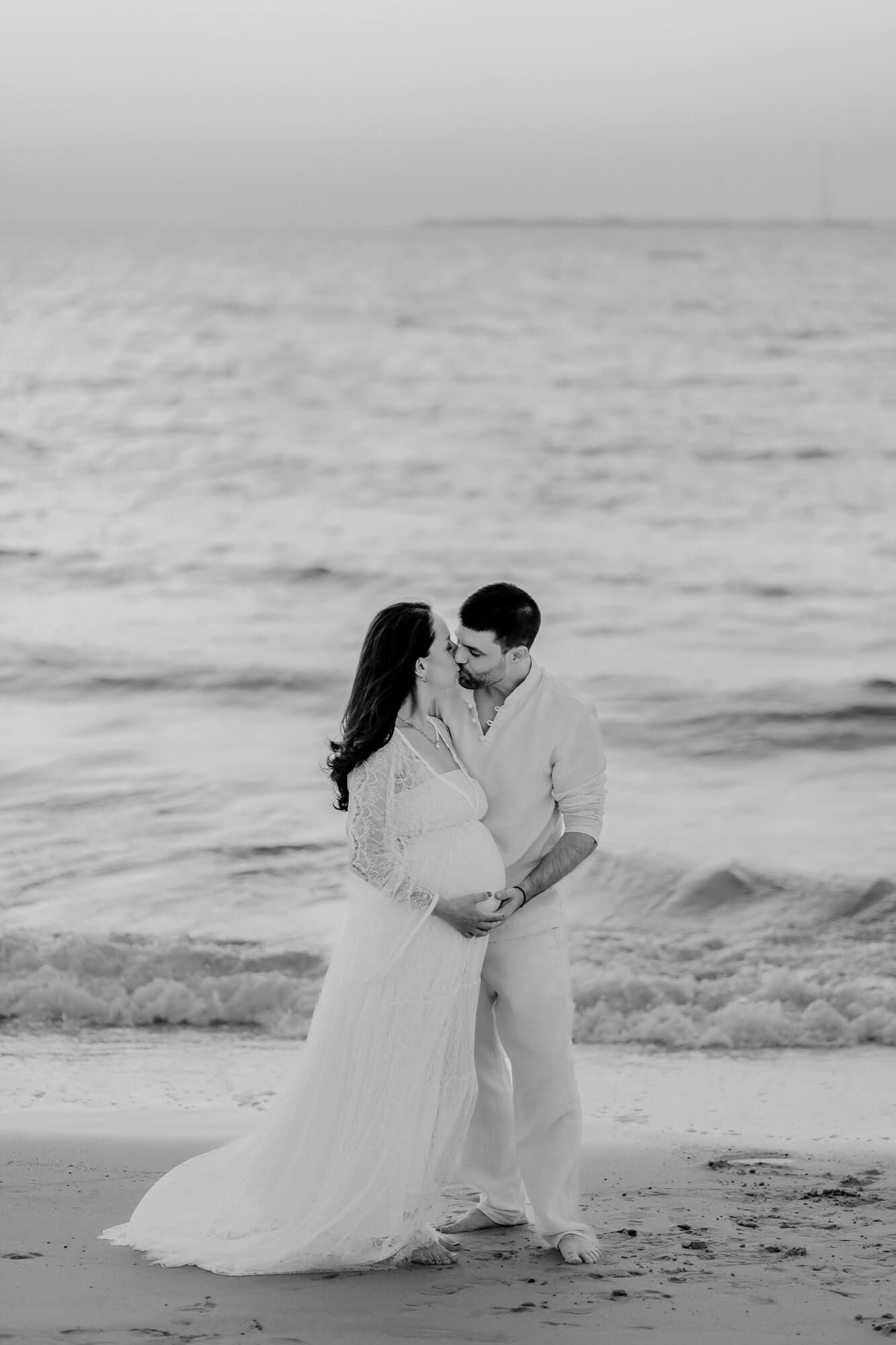 A romantic beach photo shoot featuring a couple in white attire embracing by the ocean in black and white photography.