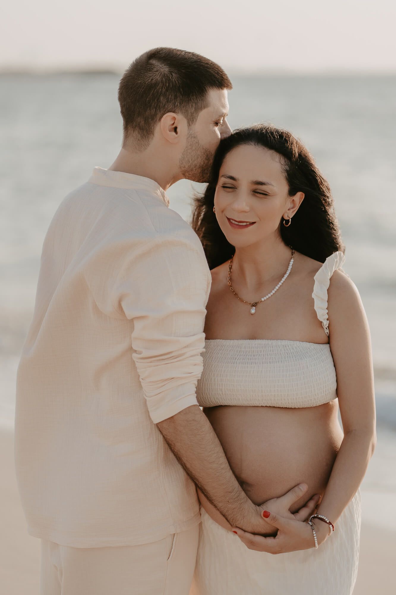 A tender maternity photo on the beach shows an expecting couple in white clothing sharing an intimate moment together.