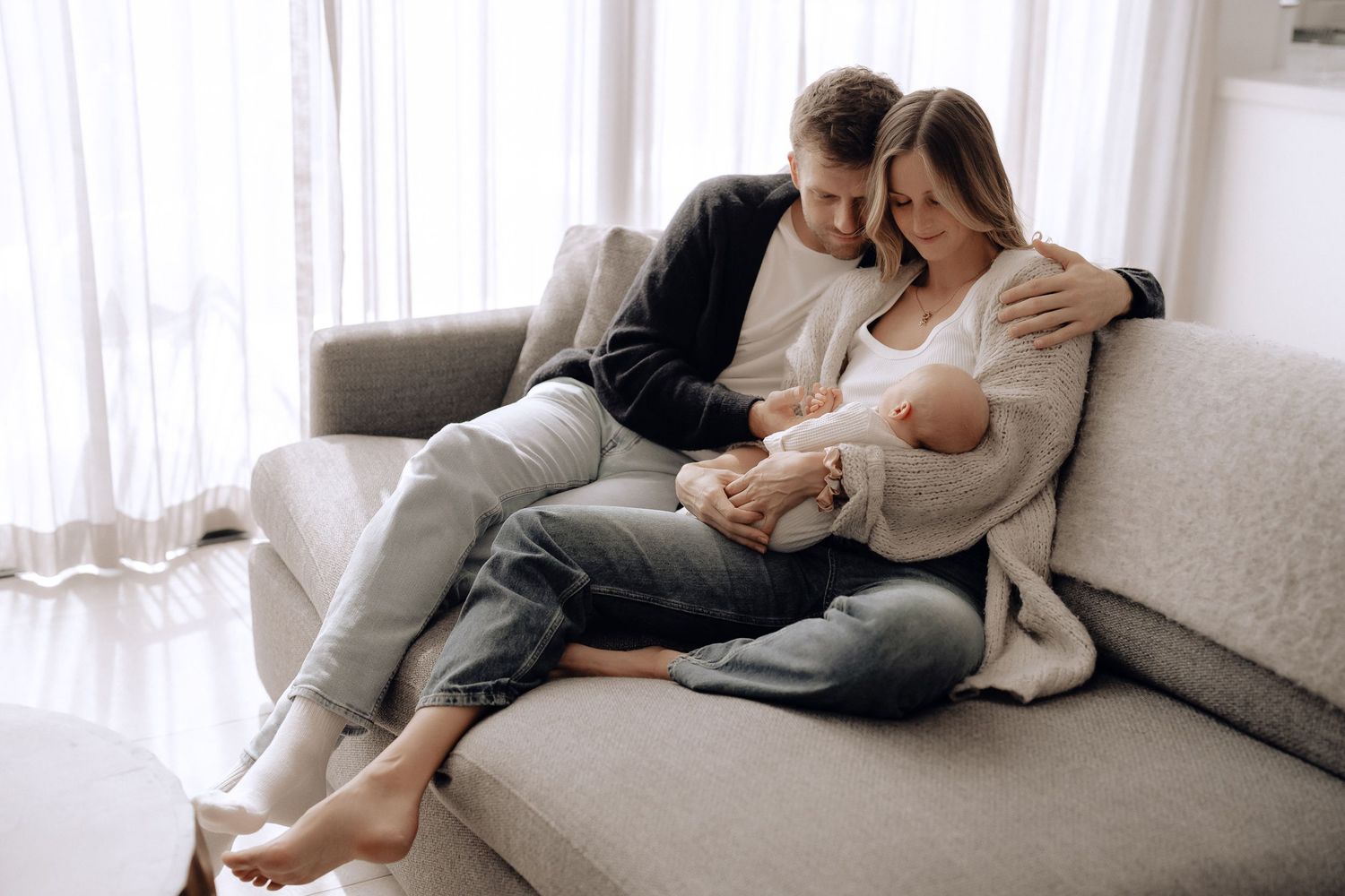 New parents sit together on a gray couch in a bright living room while holding their sleeping newborn baby.