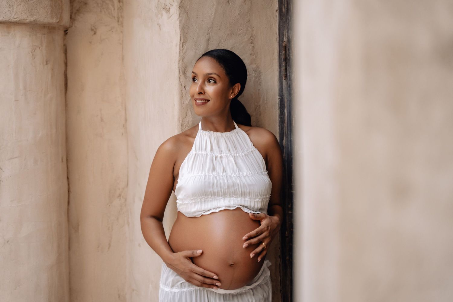 Elegant maternity portrait of expectant mother in white halter dress against textured wall.