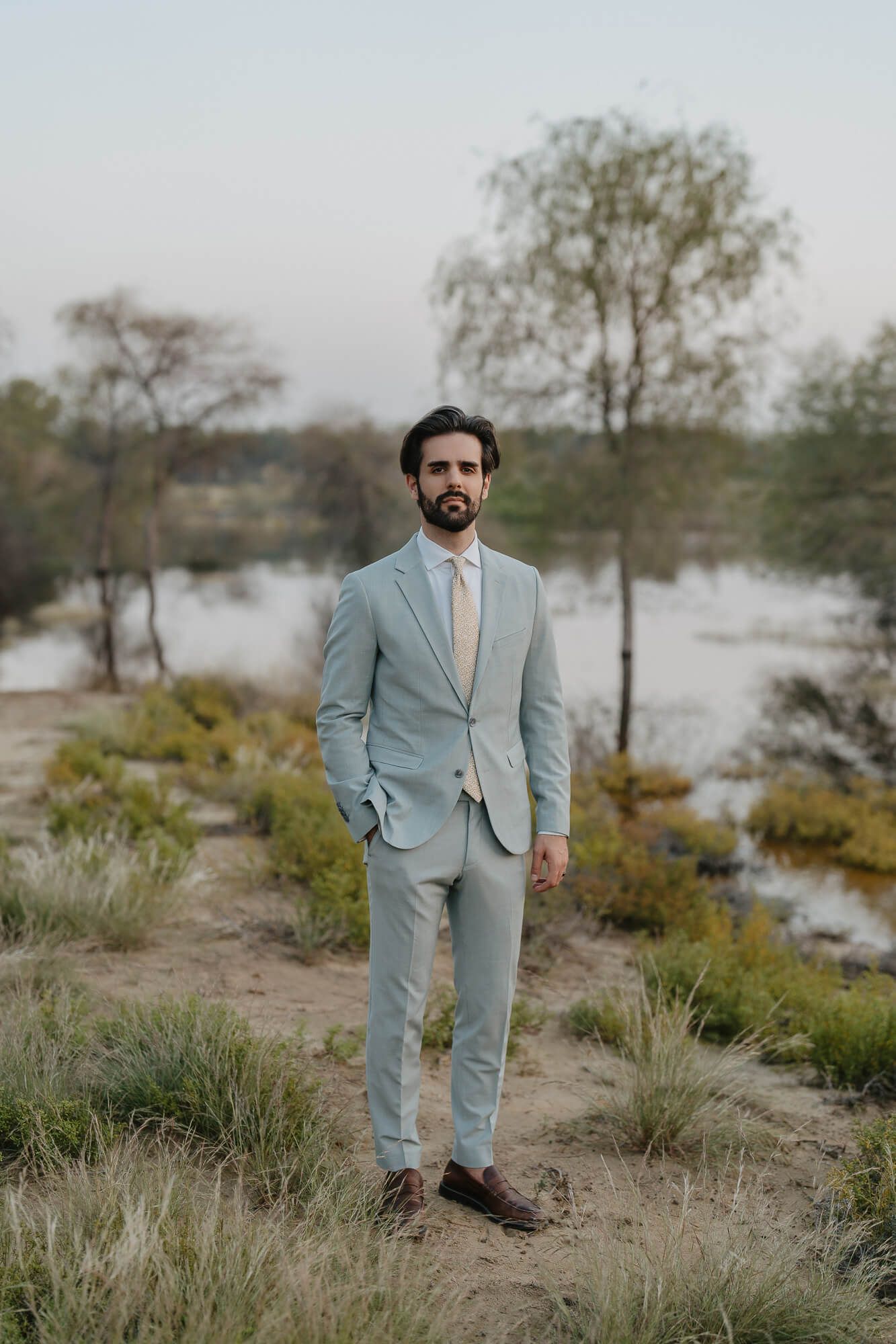 A well-dressed figure in a light blue suit stands in a desert landscape with brush and trees in the background.