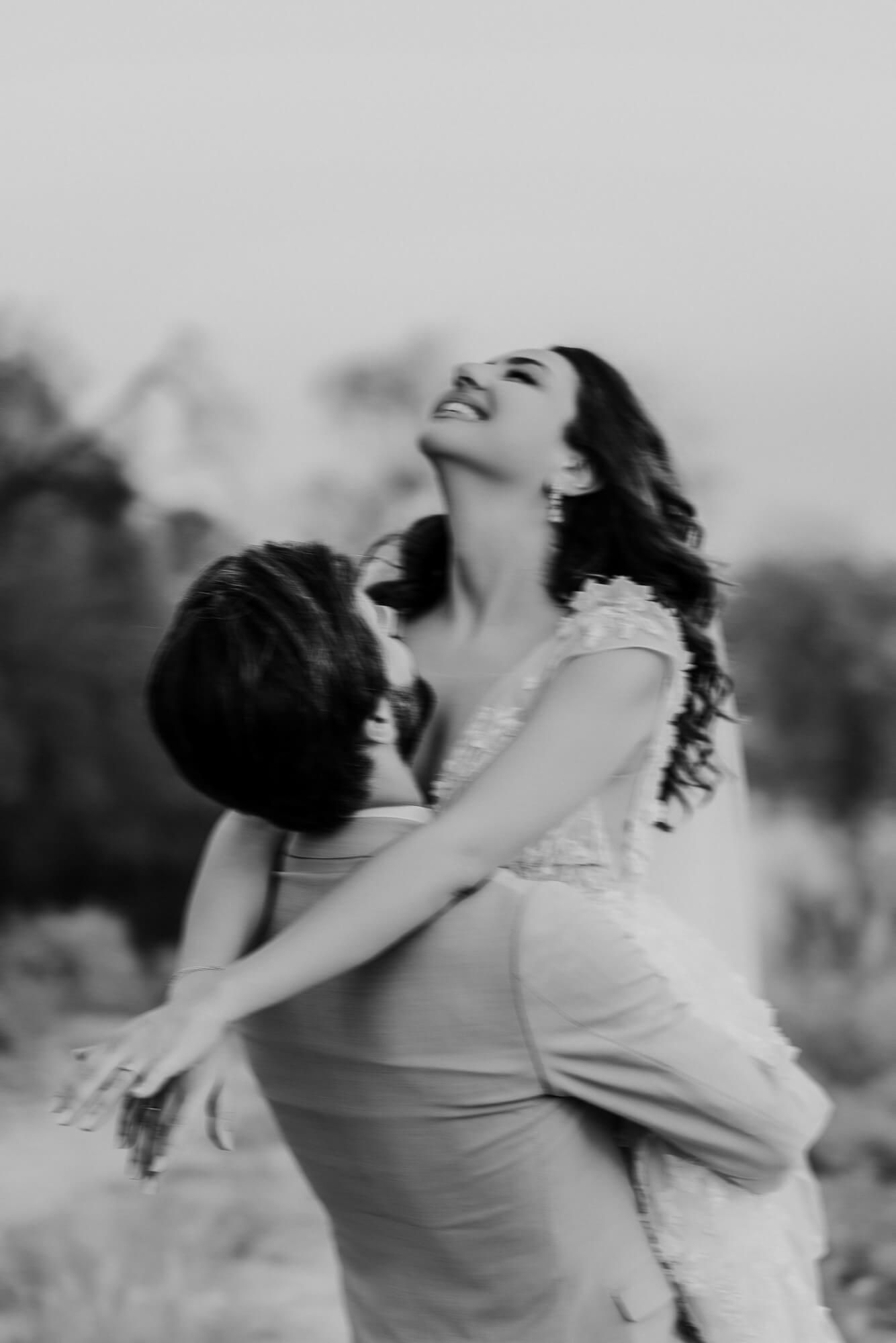 A joyful romantic black and white photo of a couple embracing outdoors with the person in a dress laughing.