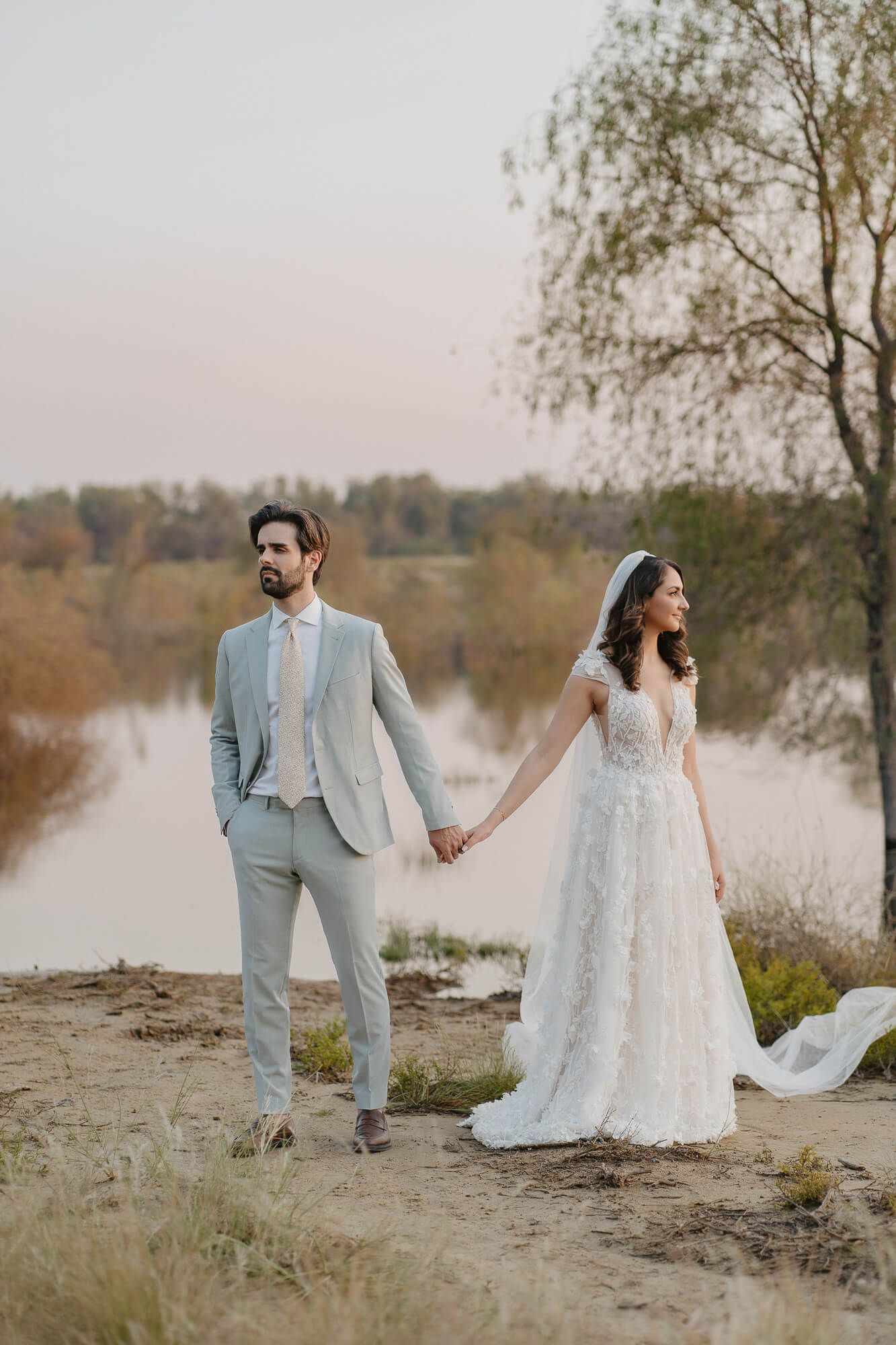 A couple in wedding attire holds hands by a scenic autumn lake with tall birch trees in the background.