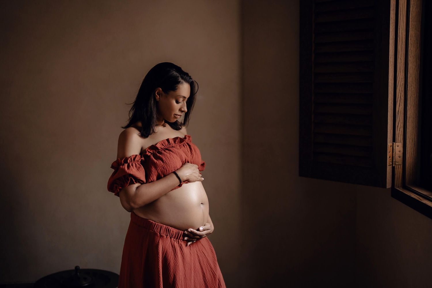An expectant mother in a red dress poses by a window in dramatic lighting.