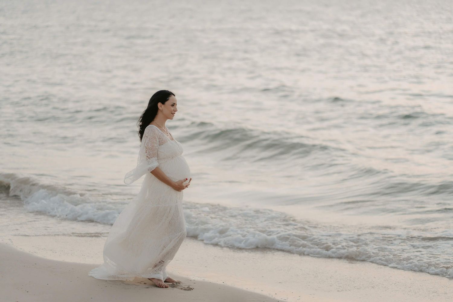 A pregnant woman in a flowing white dress stands barefoot on a beach at sunset near the ocean waves.