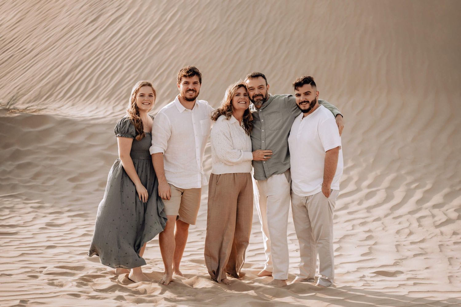 Five people in neutral tones stand together on desert sand dune during outdoor photo session.