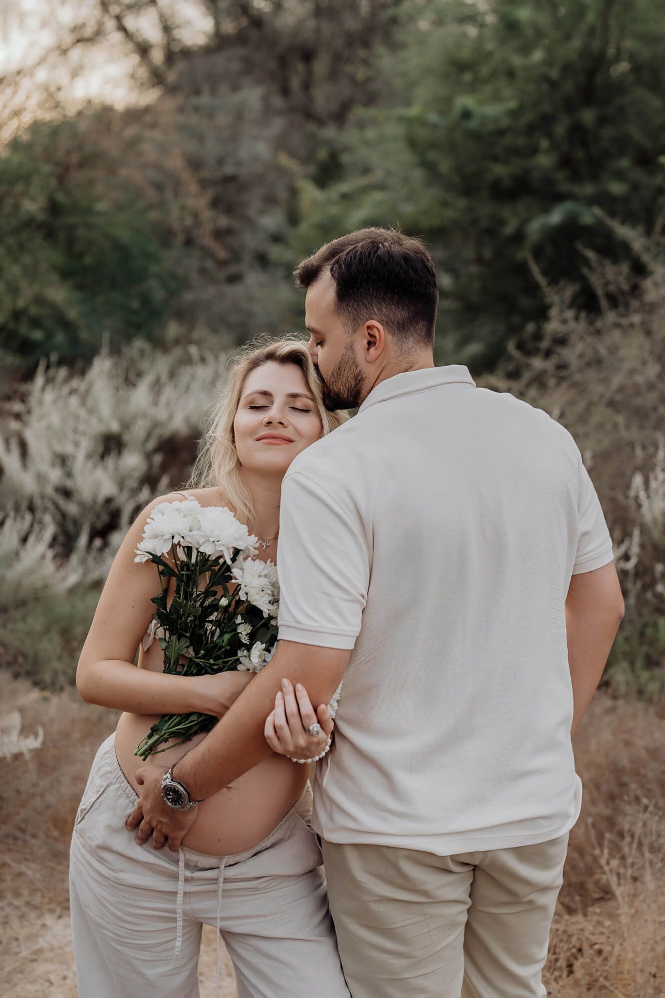 Tender maternity moment between couple dressed in white, sharing embrace with flowers in natural setting.