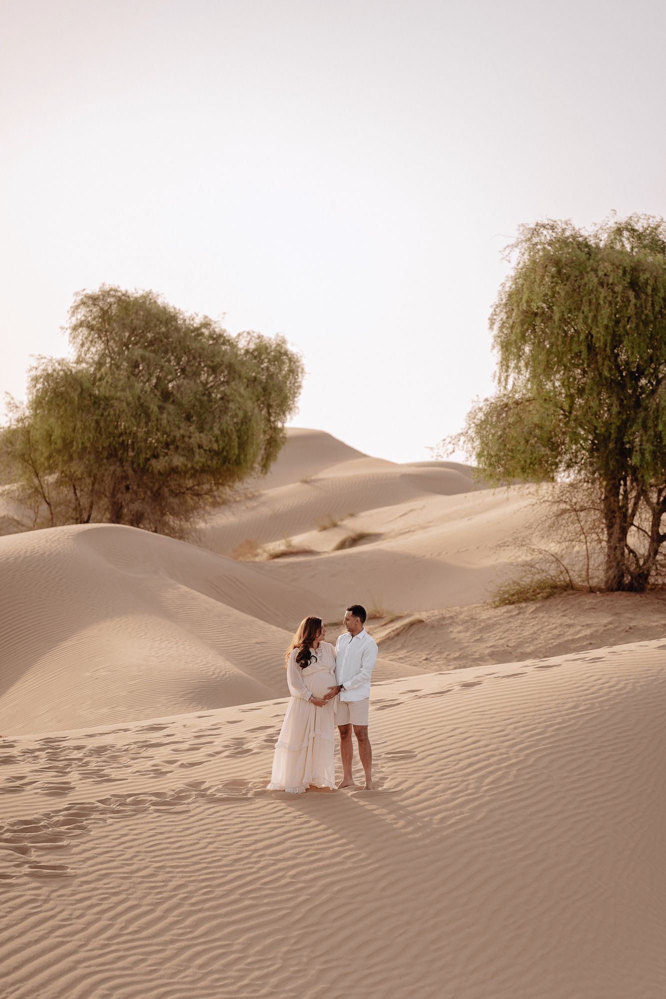Couple standing together on sand dunes with willow trees in a desert landscape at sunset.