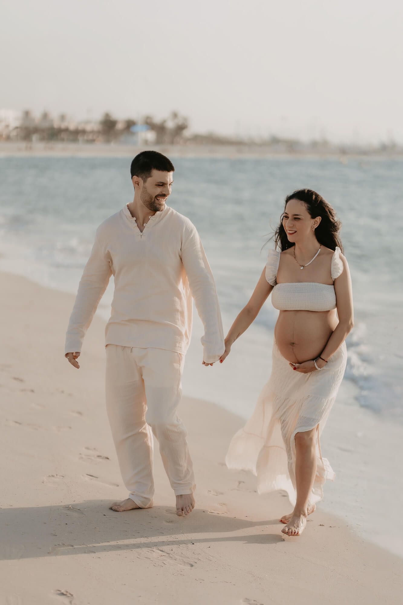 A couple wearing matching cream colored outfits walk hand in hand along a sandy beach during a romantic photoshoot.