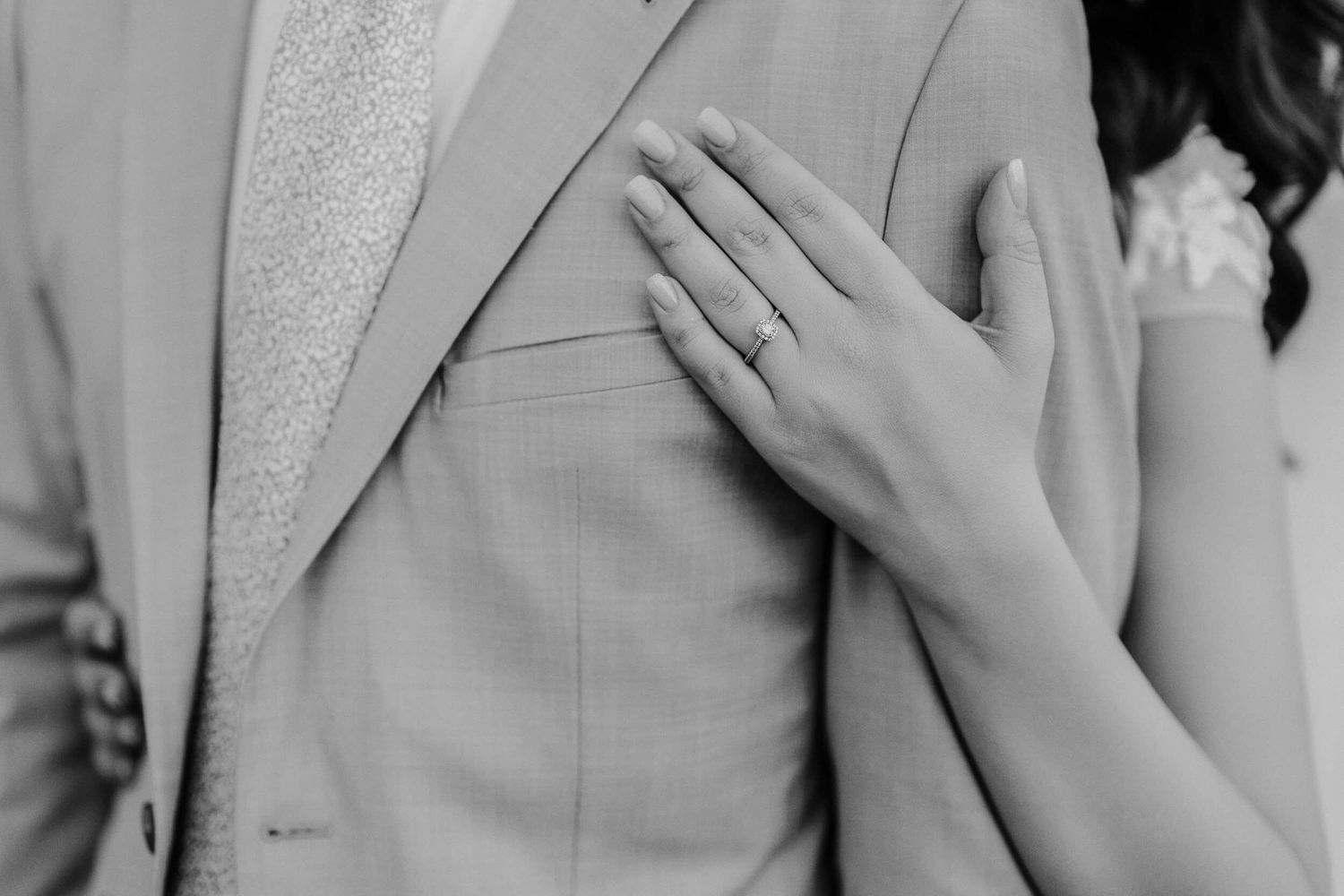 Close-up black and white photo showing bride's hand resting on groom's light colored suit jacket.