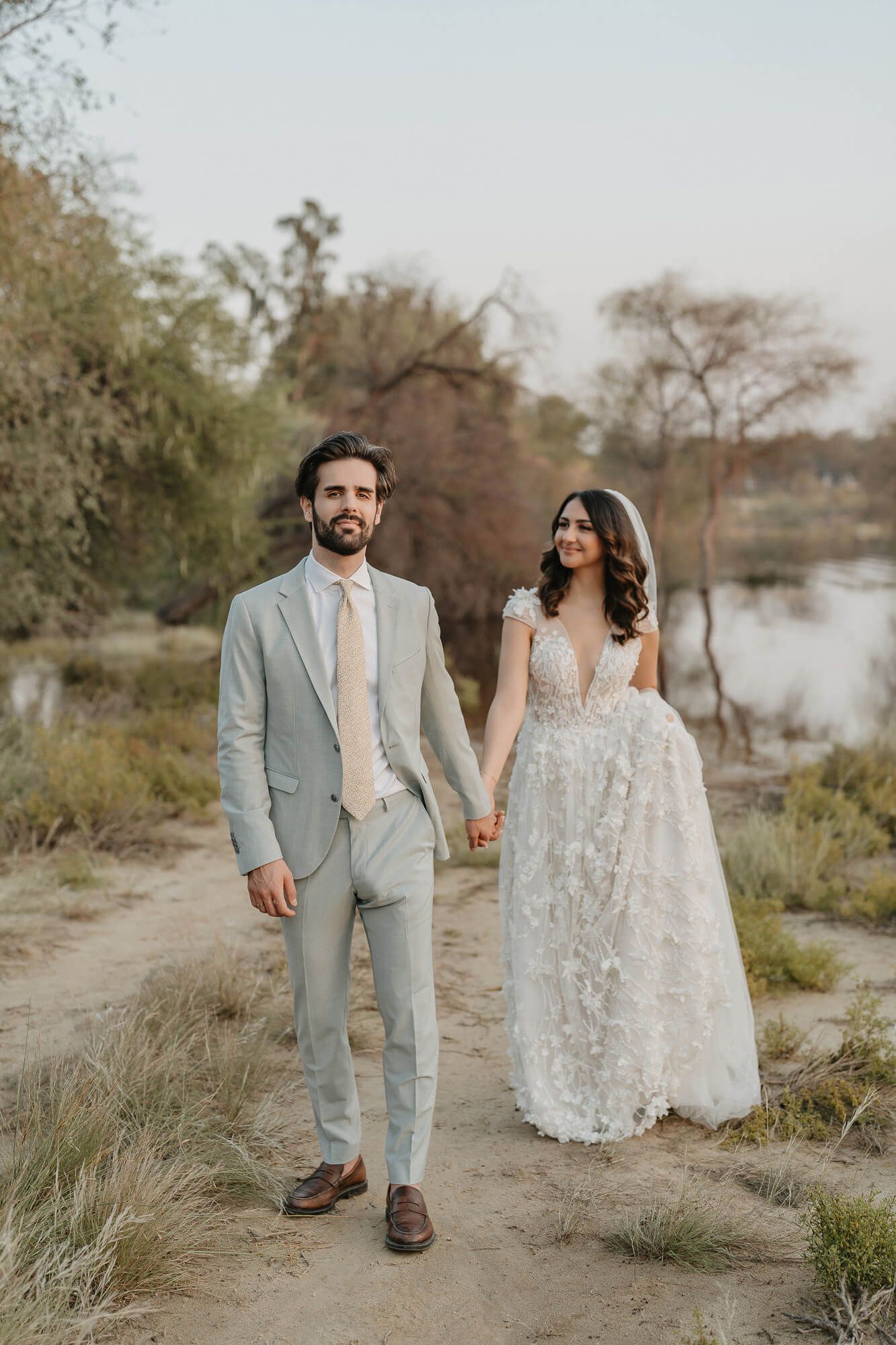 A couple in wedding attire walk hand in hand along a riverbank path during a romantic outdoor photoshoot.
