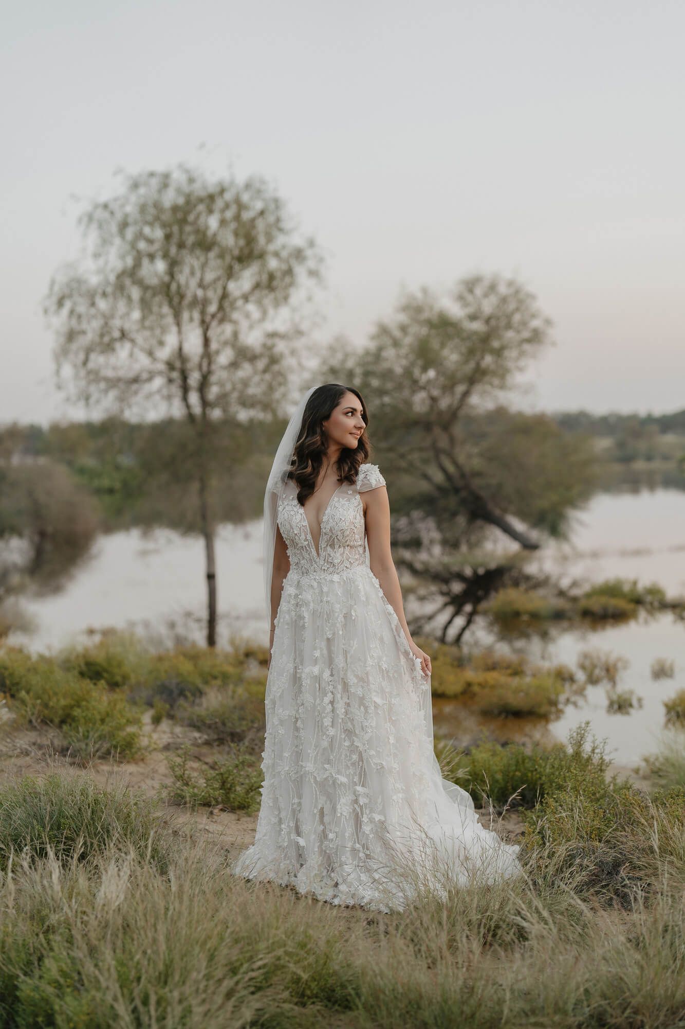 A bride in a detailed lace wedding gown stands on a grassy hillside overlooking a serene lake at dusk.