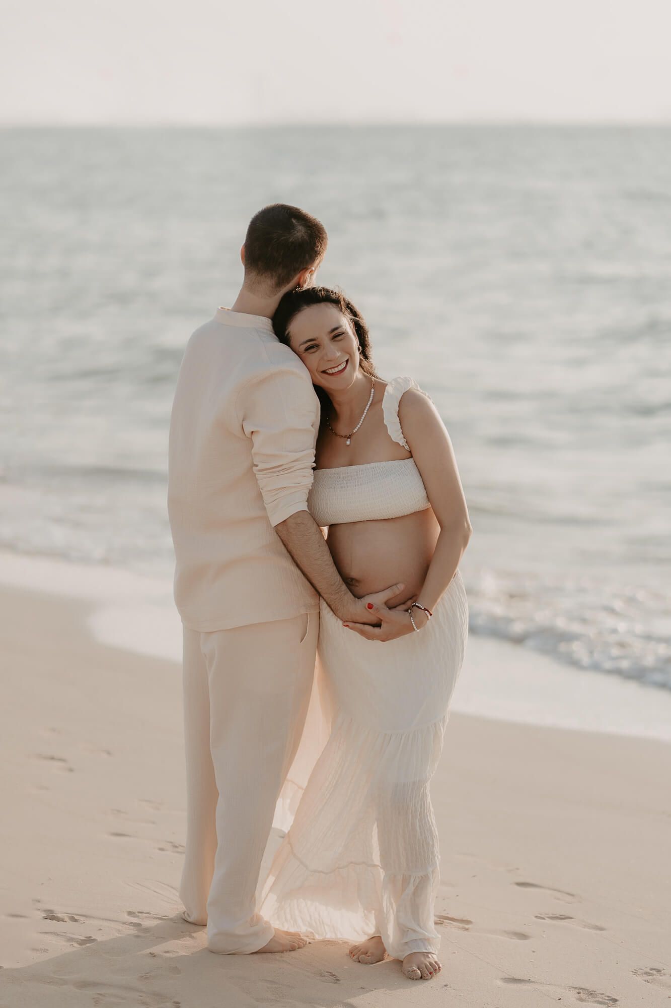 Romantic maternity photoshoot on the beach with couple in matching white outfits at sunset.