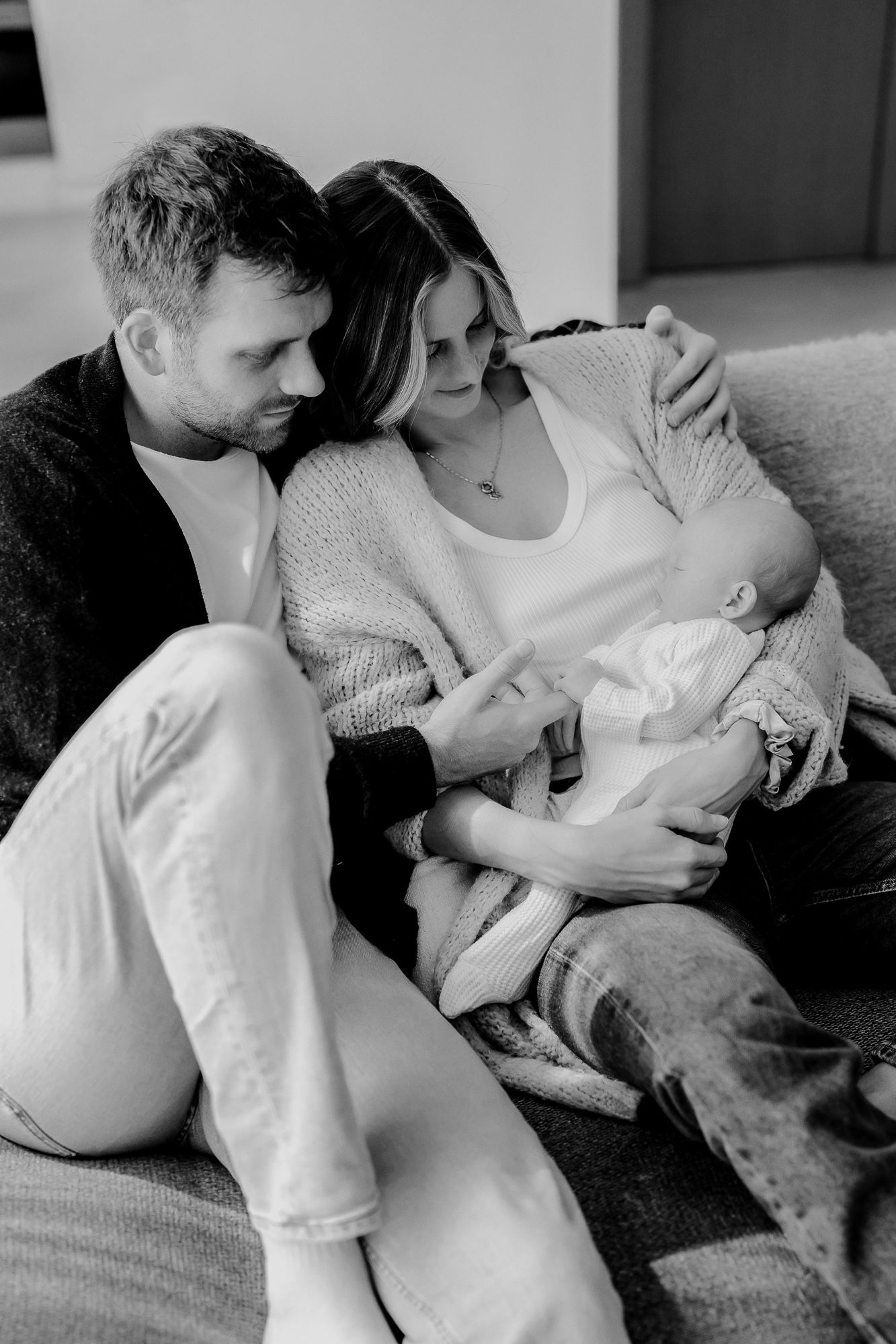 Black and white photo of a couple sitting close together on a couch holding a baby in a peaceful, tender moment.