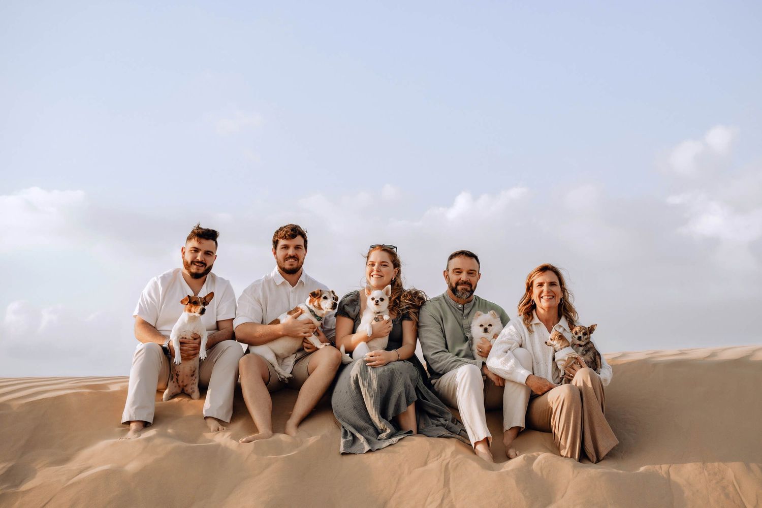 Family group seated on sand dunes holding small dogs during a sunset photoshoot.