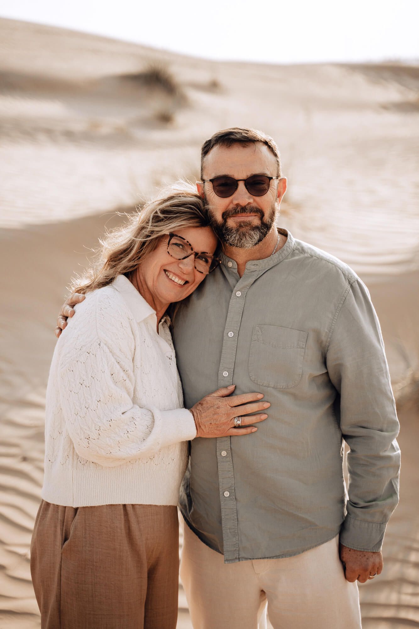 Couple in casual attire embracing in front of sand dunes during golden hour.