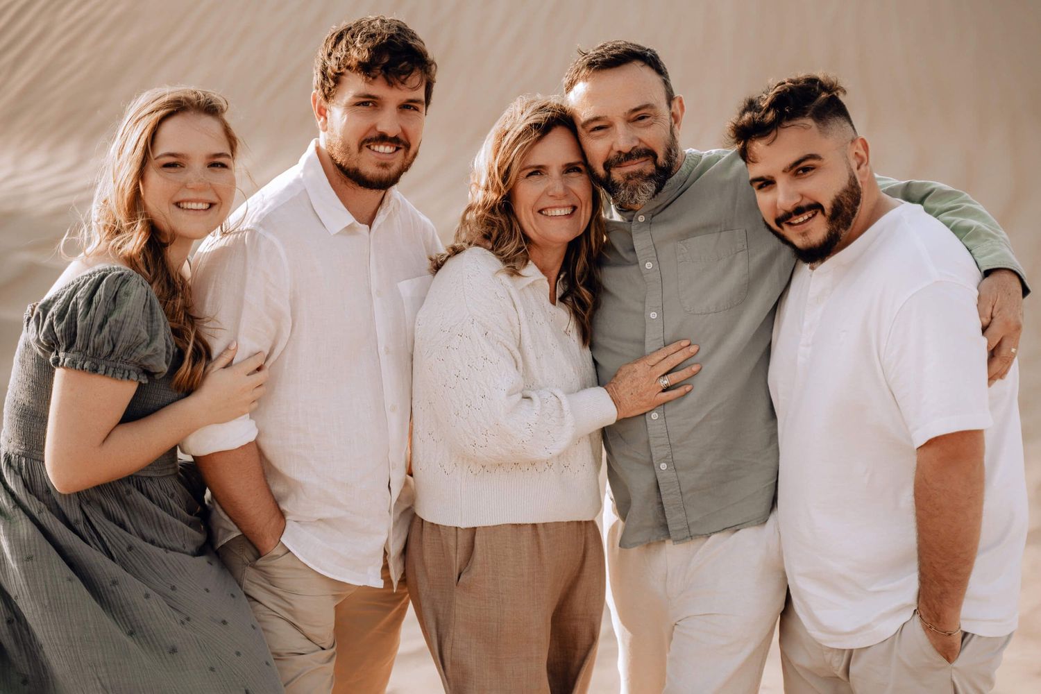 Family group photo of five people in coordinated casual outfits posing together against desert sand dunes.
