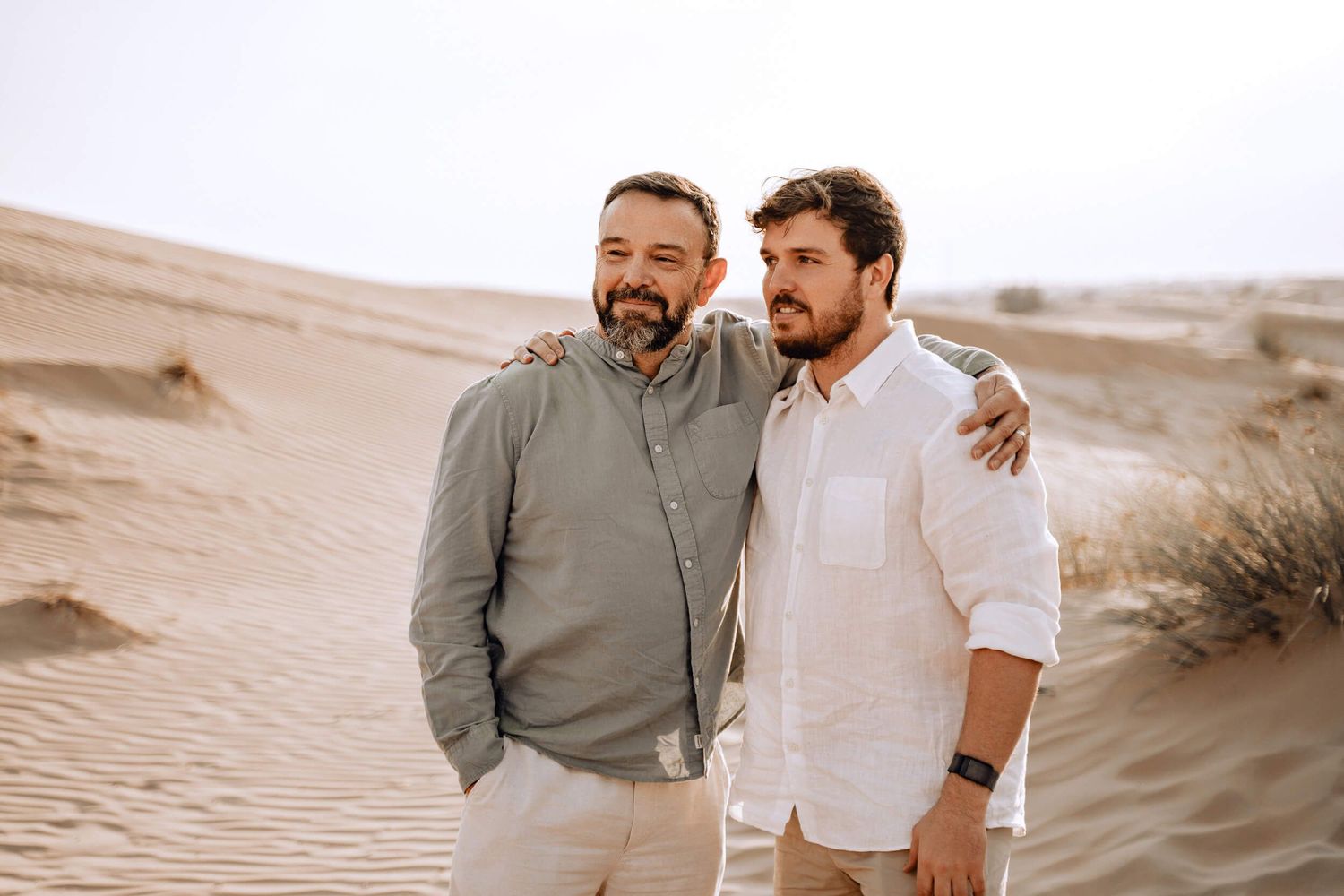 Two people smile and embrace while standing on desert sand dunes in casual attire.
