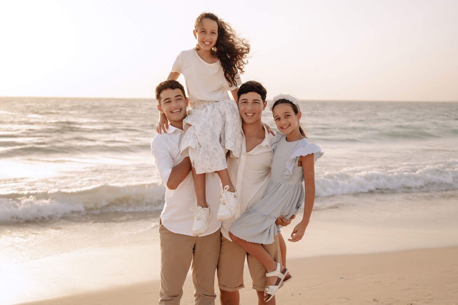 Family wearing coordinated white and beige outfits smiling together on sandy beach.