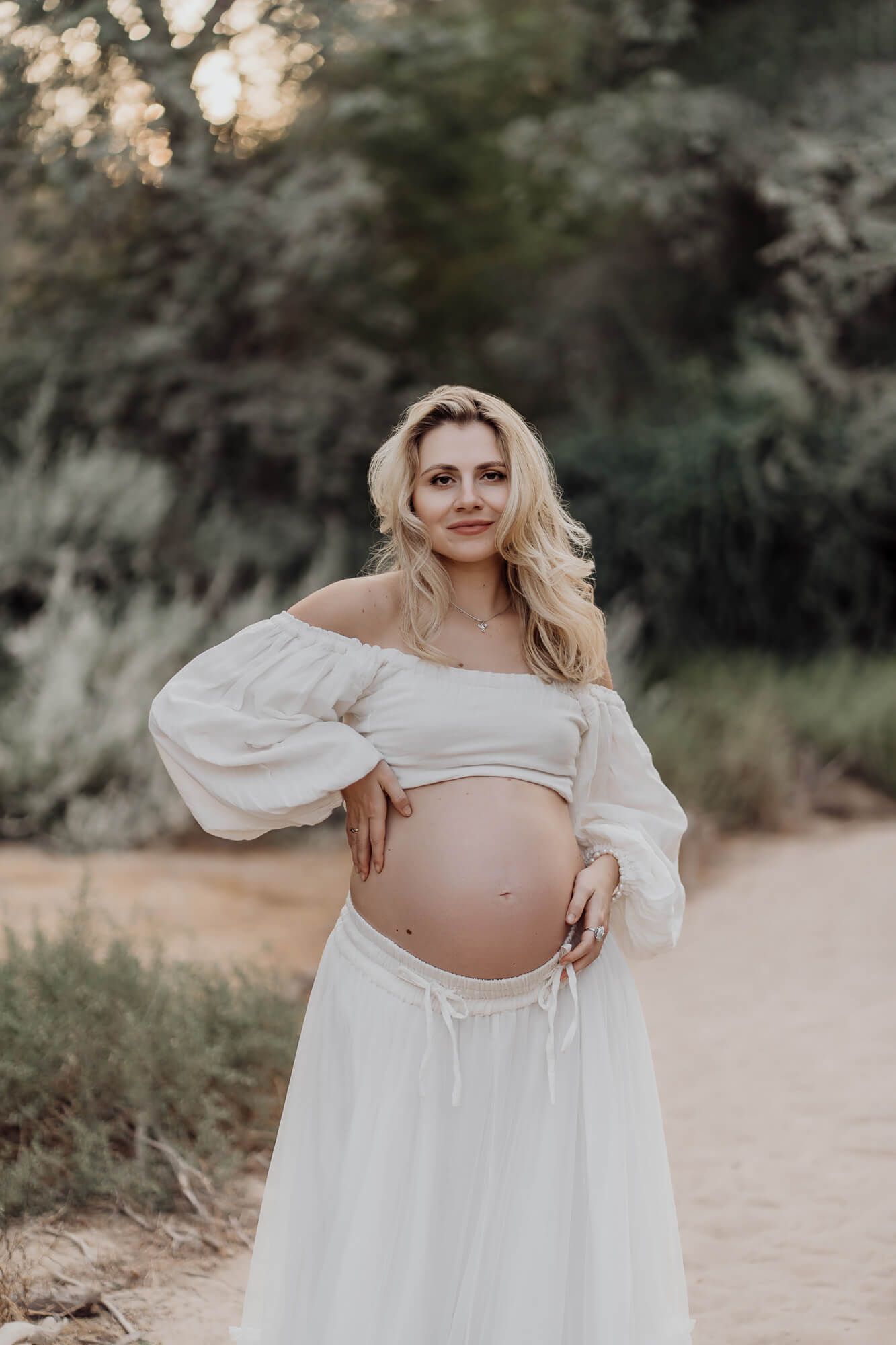 Maternity portrait of woman in white dress showcasing baby bump against natural backdrop.