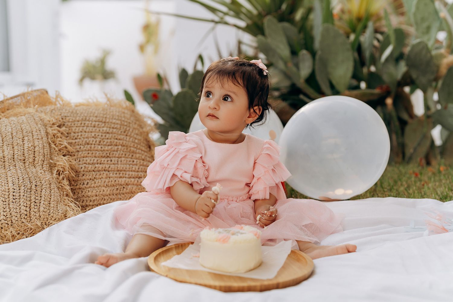 A baby in pink ruffled dress sitting beside balloon and woven basket on white blanket. Cake smash shoot