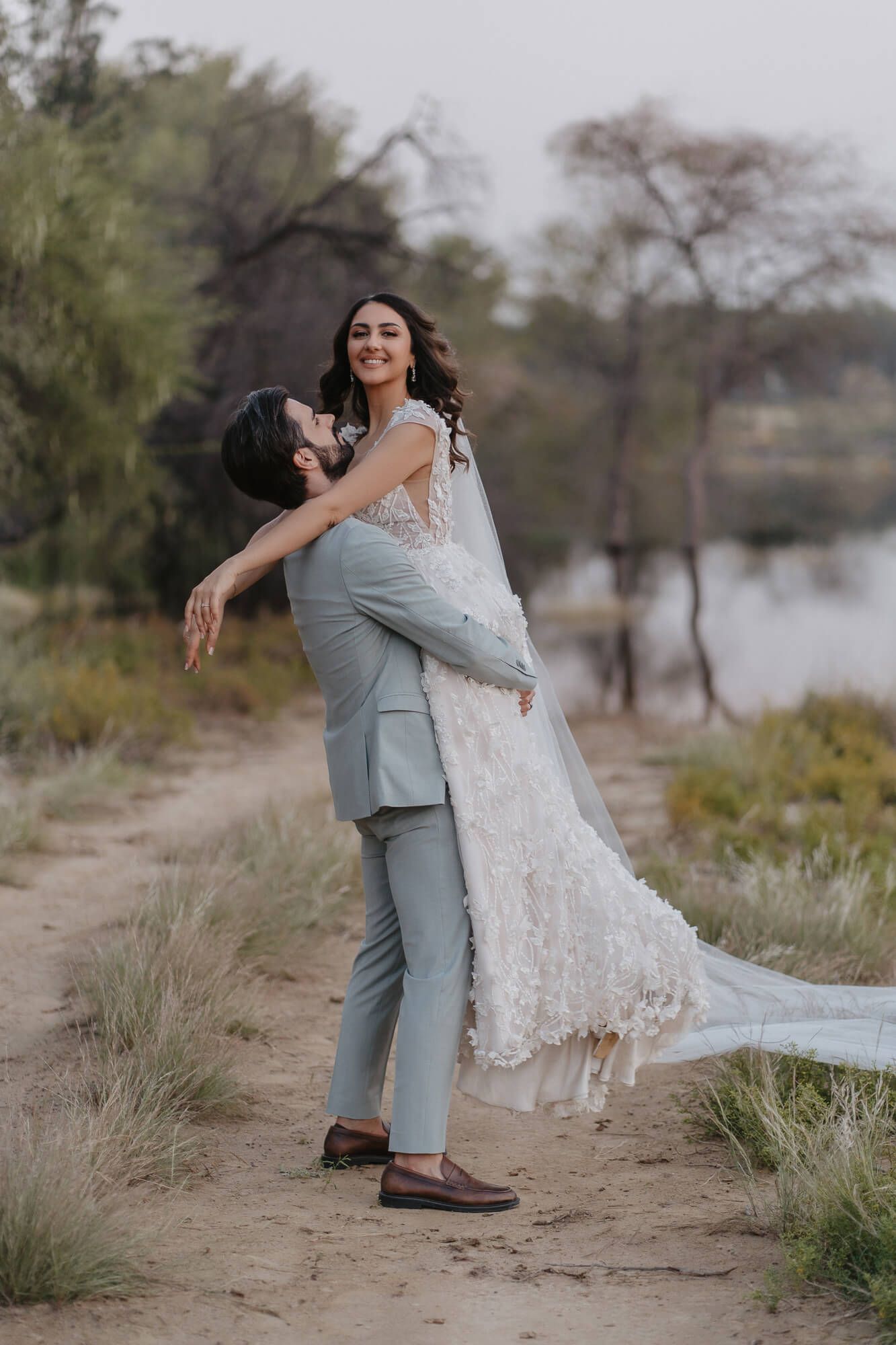 A couple in wedding attire embrace joyfully on a dirt path near a lake with trees in the background.