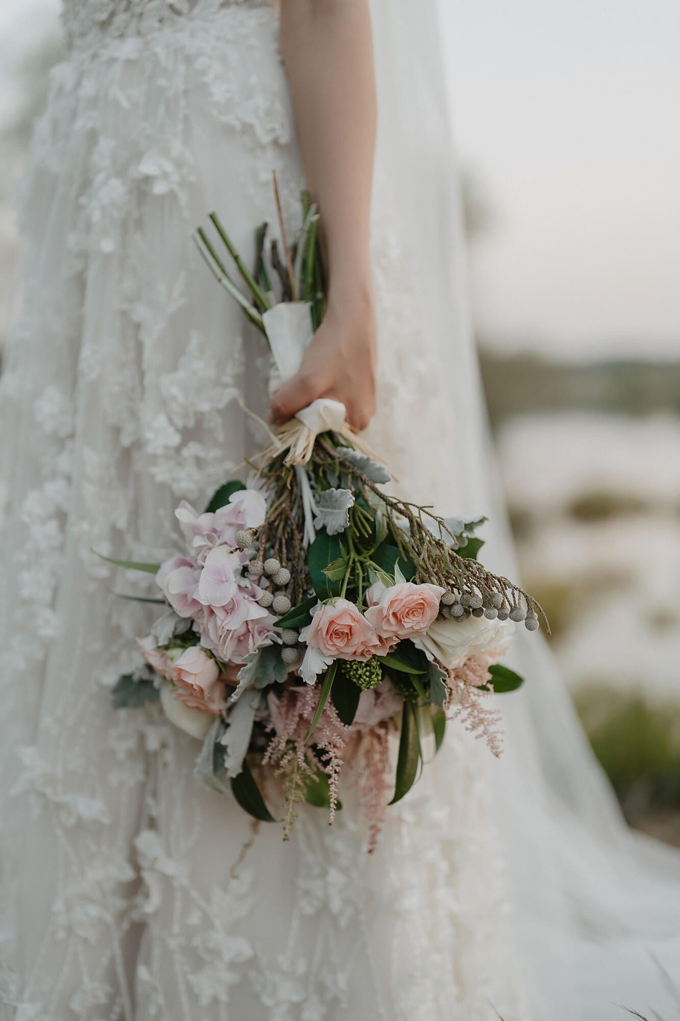 A delicate bridal bouquet of pink roses and flowers is held against a white lace wedding dress in soft lighting.
