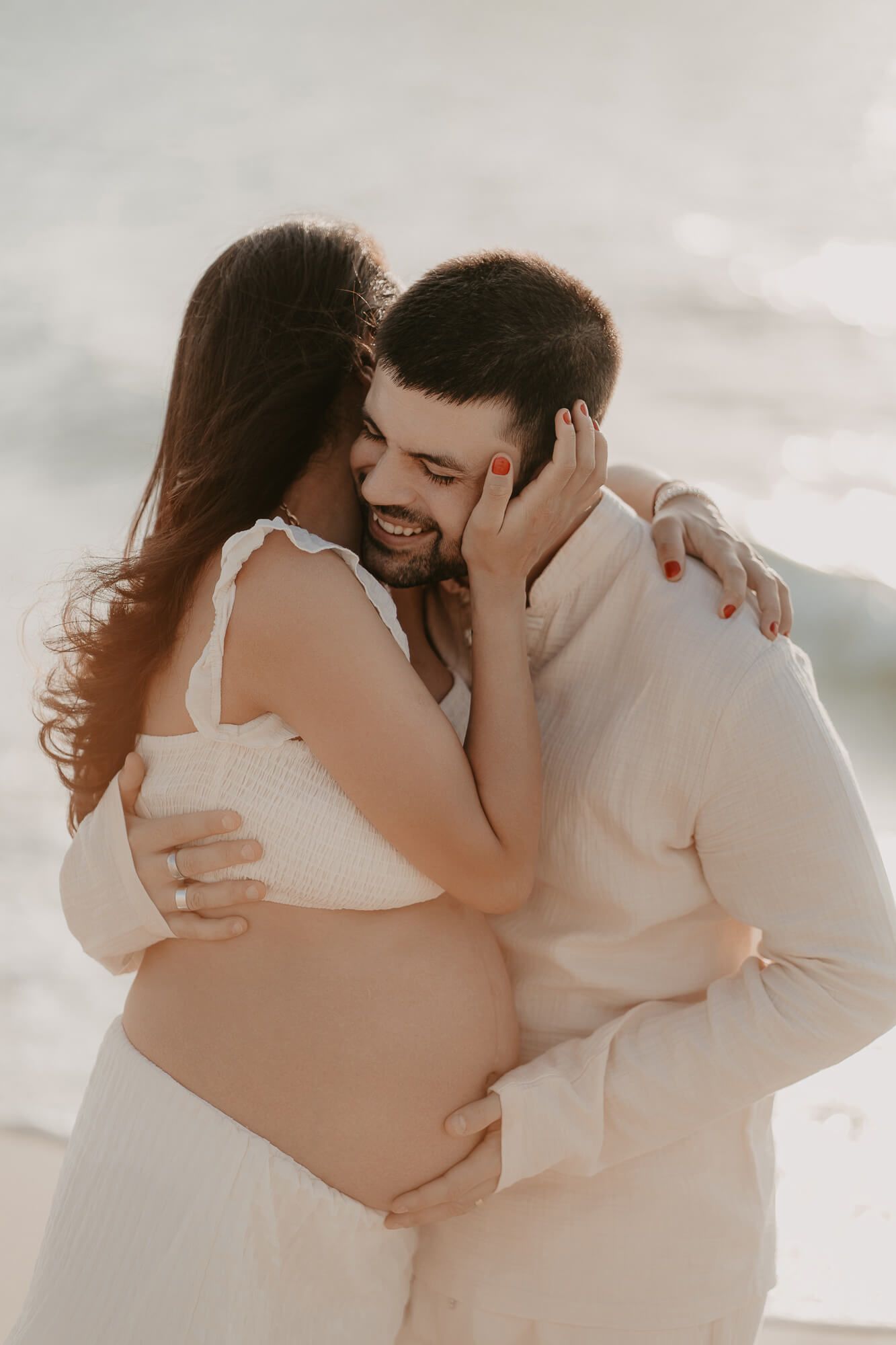 An intimate maternity photo shows a tender moment between a couple in white clothing embracing outdoors during sunset.