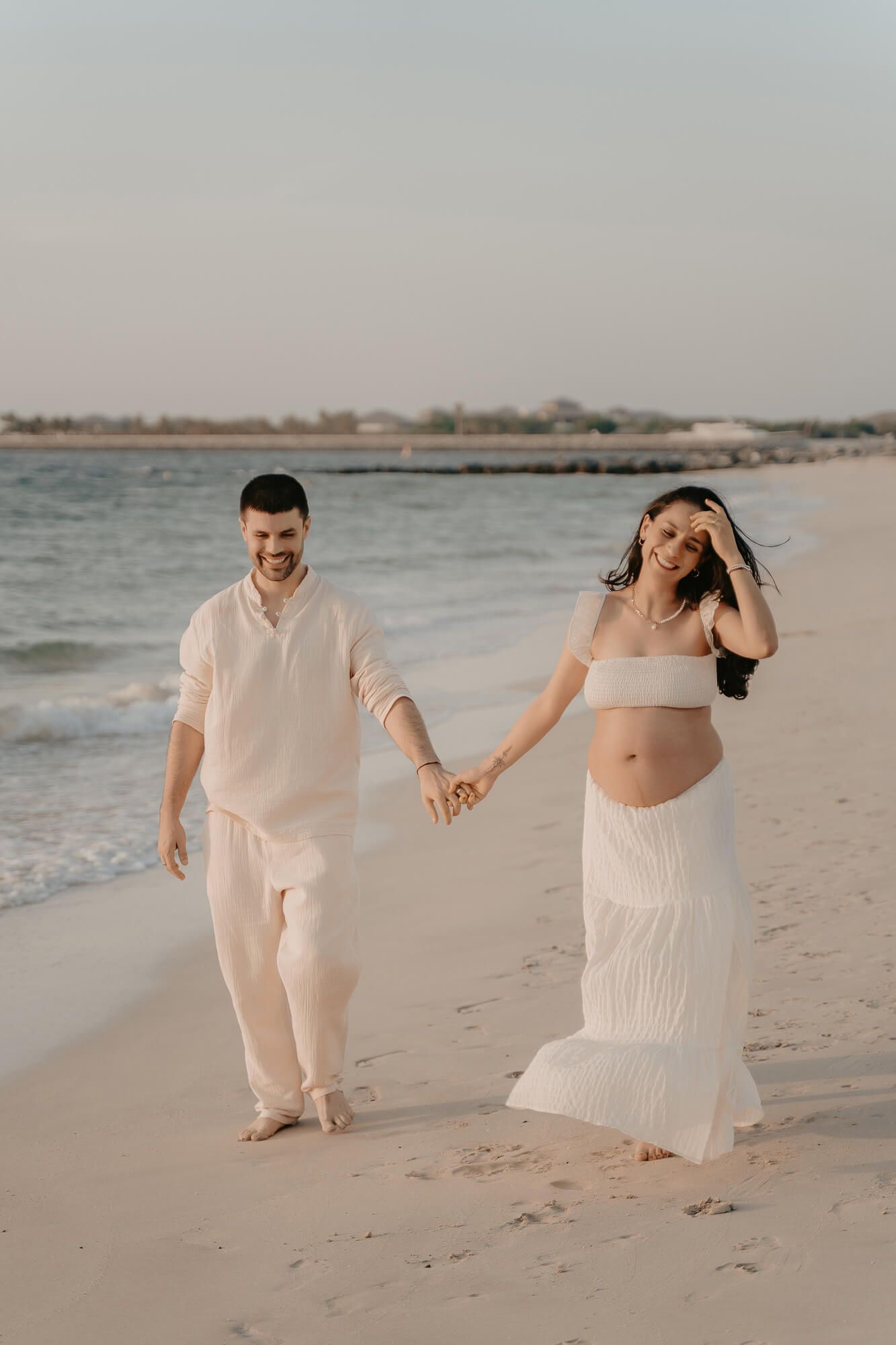 A pregnant couple in white outfits holding hands while walking on a beach during an overcast day.