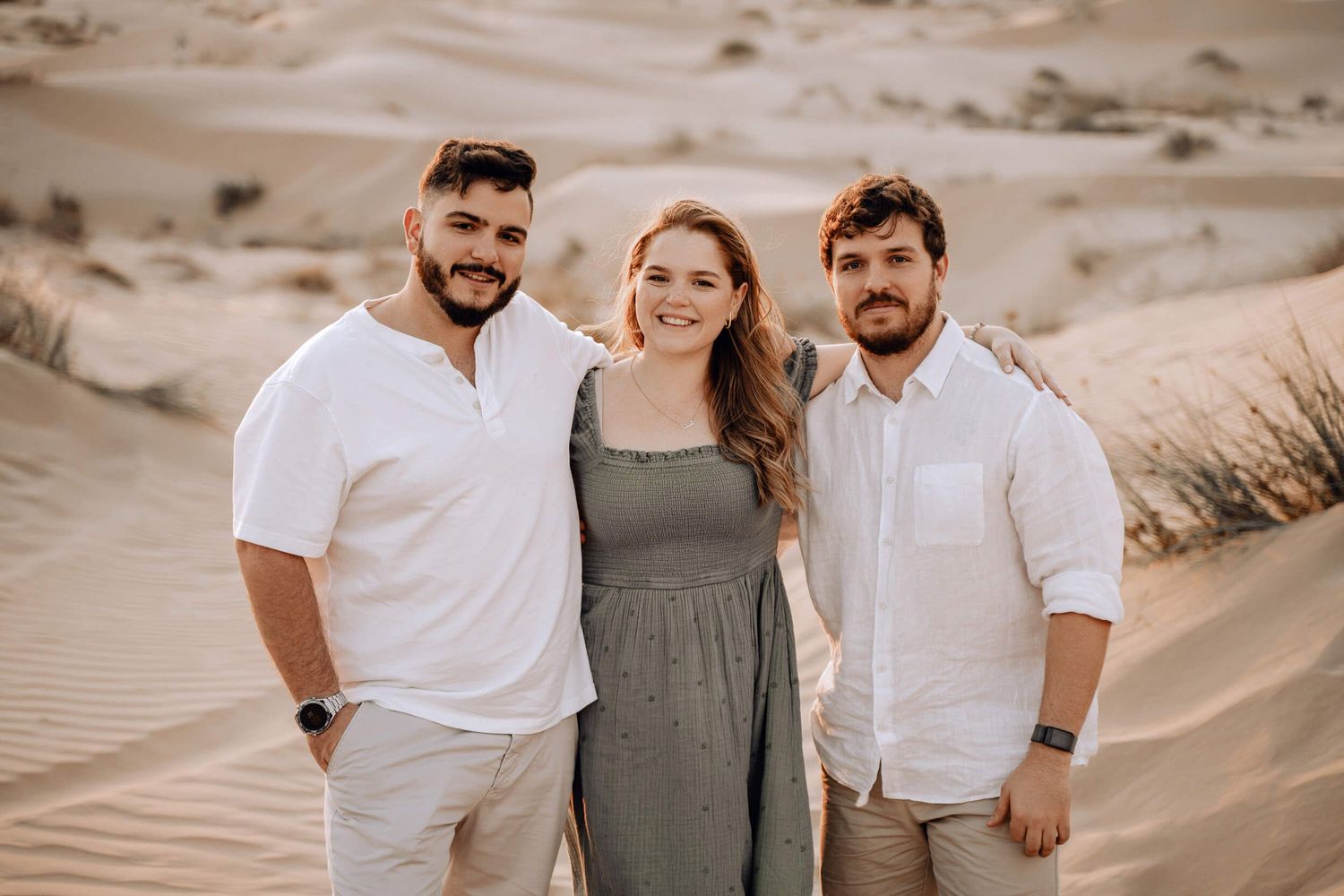 Three friends pose together for a photo in the desert sand dunes wearing coordinated white and neutral outfits.
