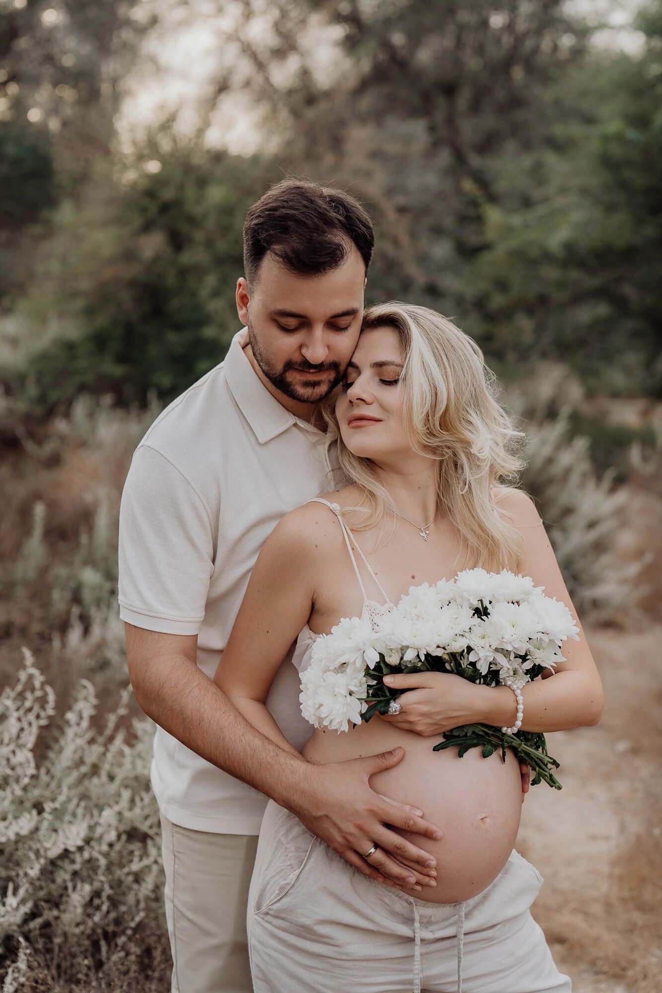 Romantic maternity portrait with couple in white clothing holding flowers in a garden setting at dusk.