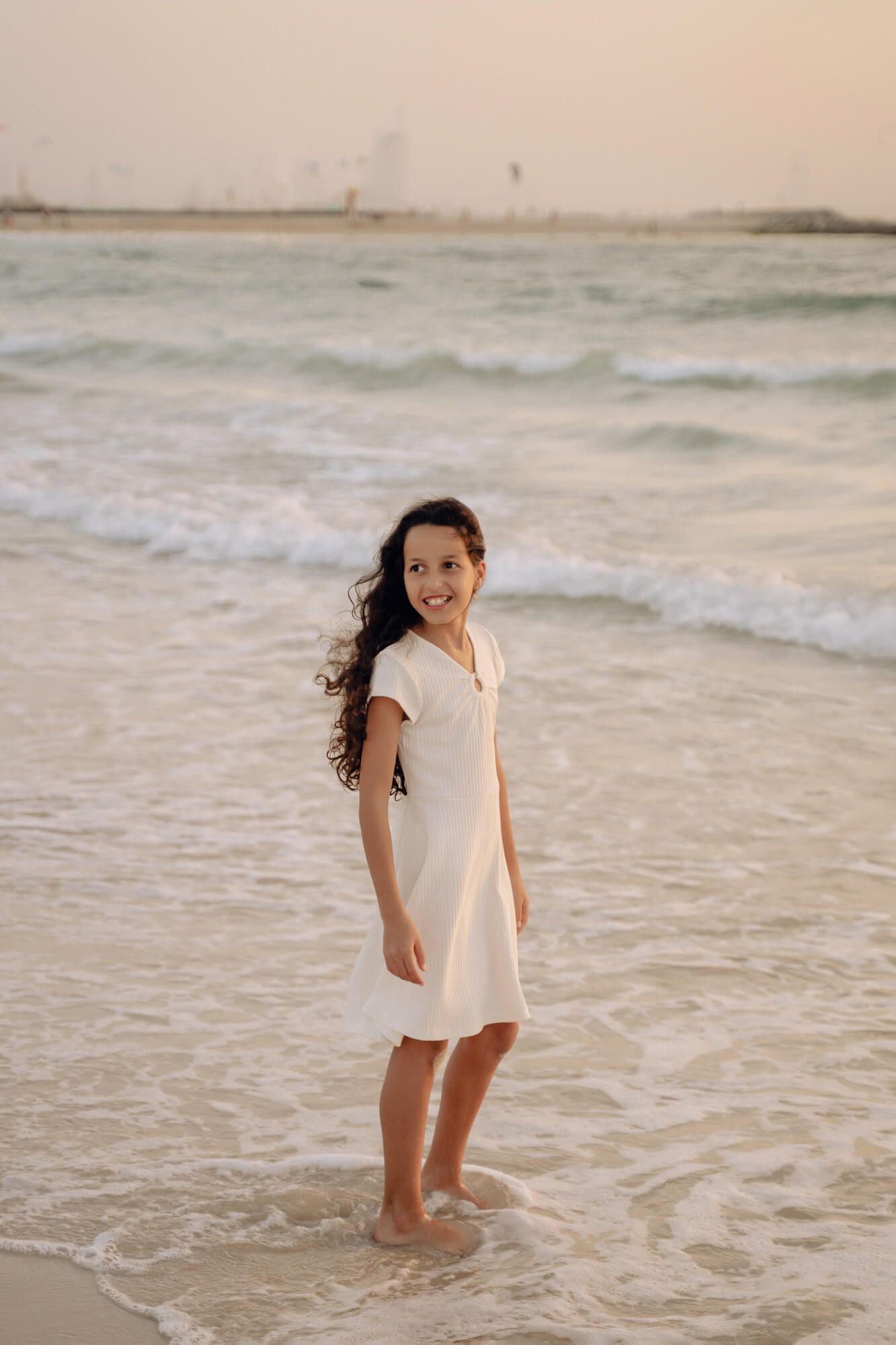 A person in a casual white dress stands smiling at the beach during golden hour.