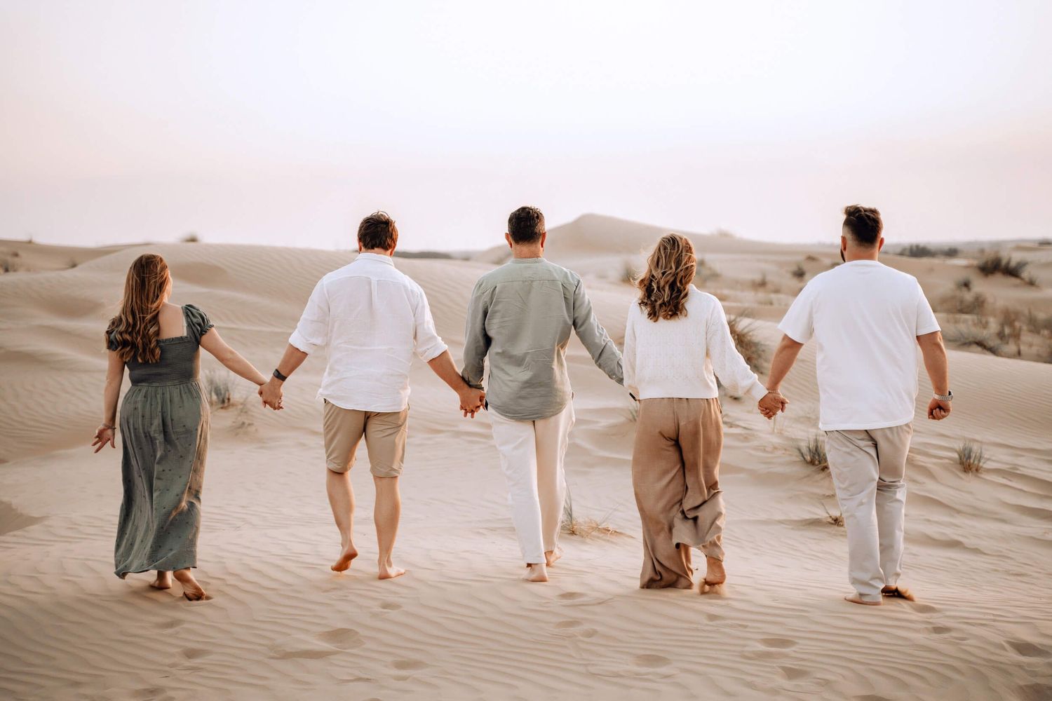 Group of five friends walk hand in hand across desert sand dunes in coordinated outfits.