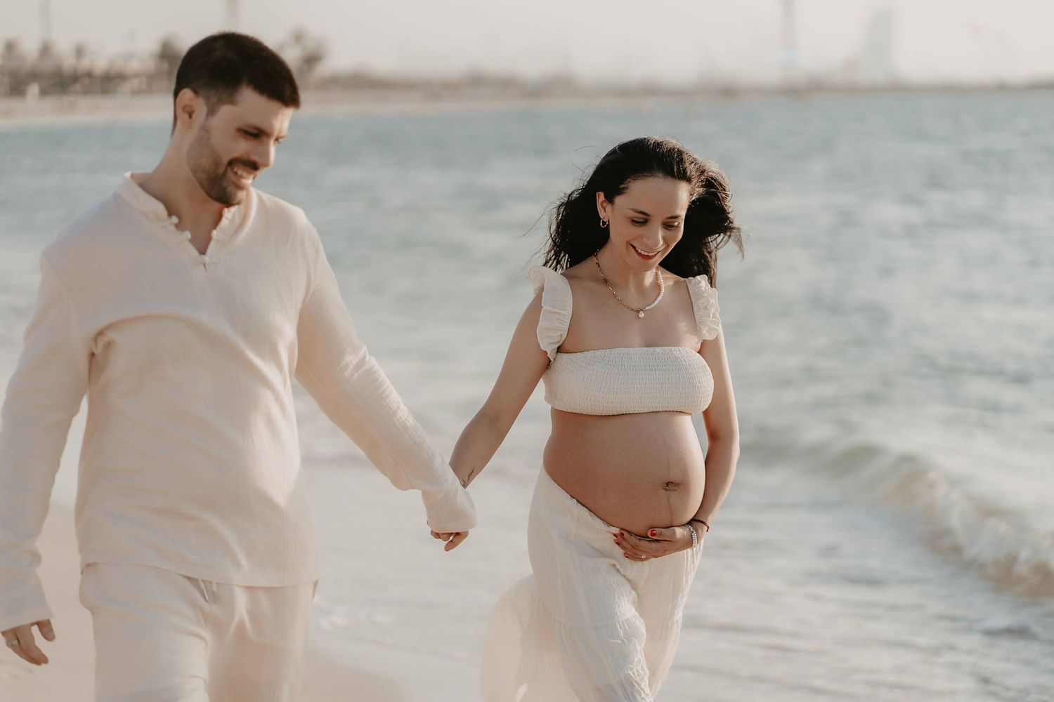 A couple wearing white outfits walks hand in hand along a beach during a maternity photoshoot at sunset.
