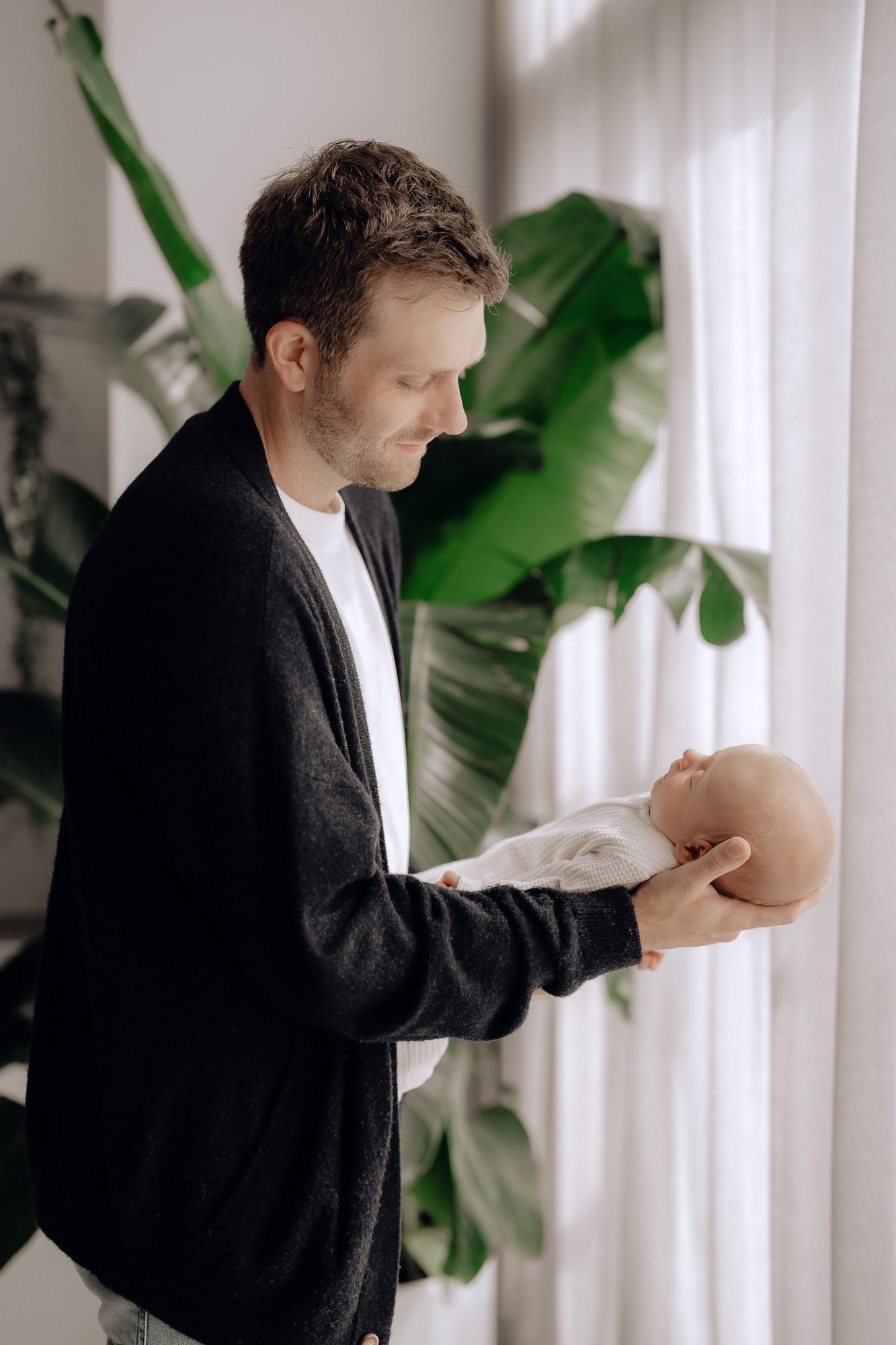 Person in black cardigan holding baby by window with large green plant in background.