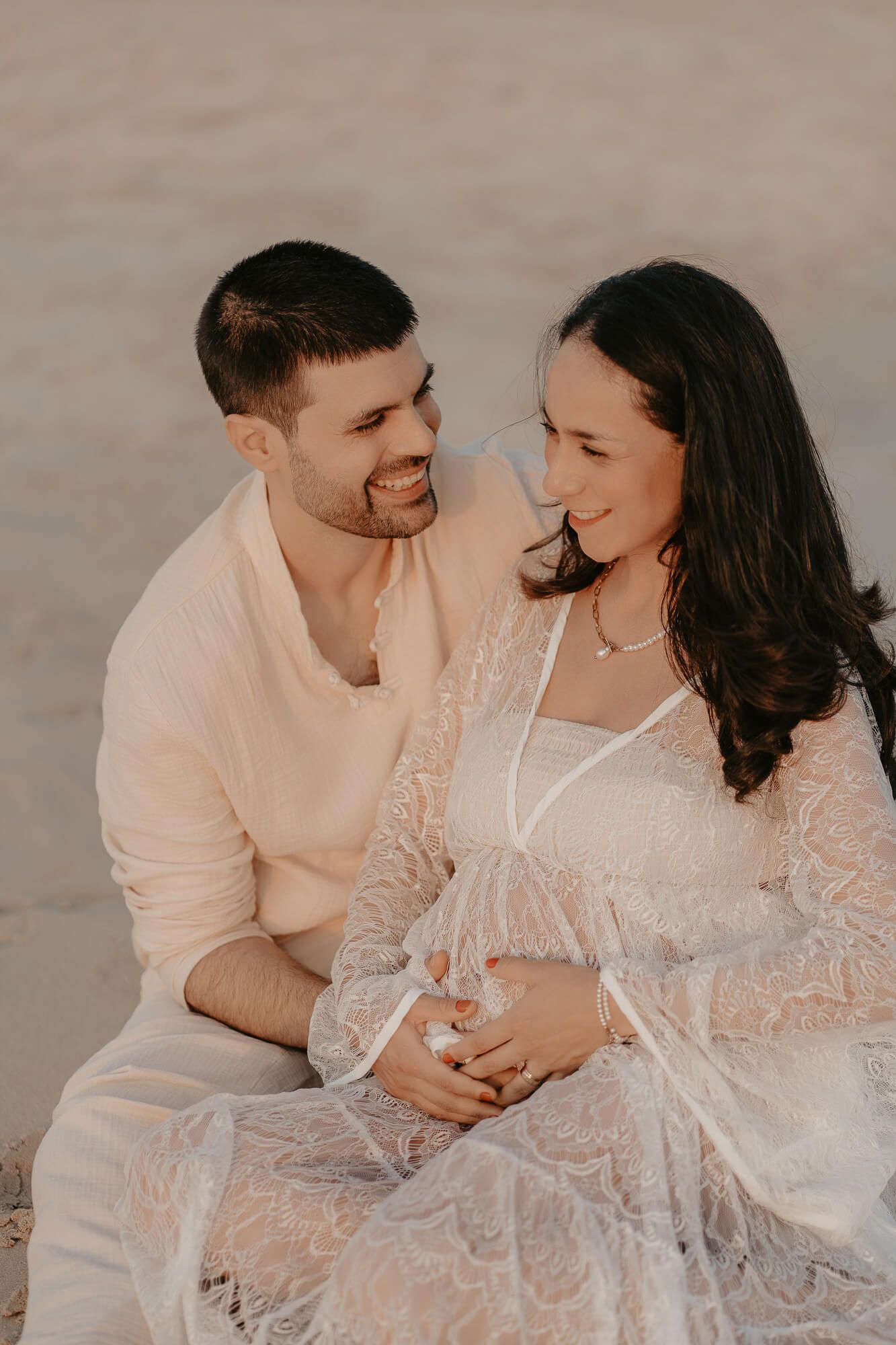 Intimate maternity photo series showing couple in white lace dress and shirt embracing on sandy beach at sunset.