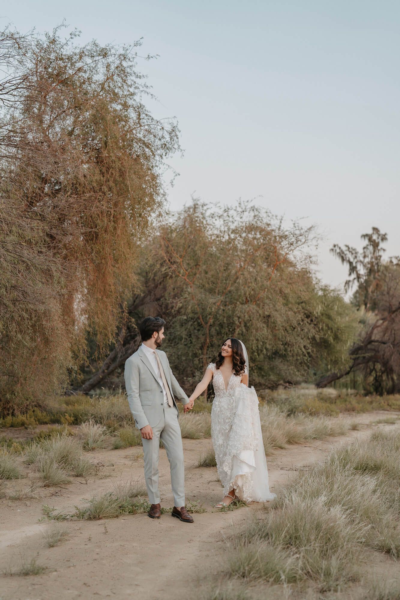 Two people in wedding attire hold hands while walking on a desert path with willow trees in the background at sunset.