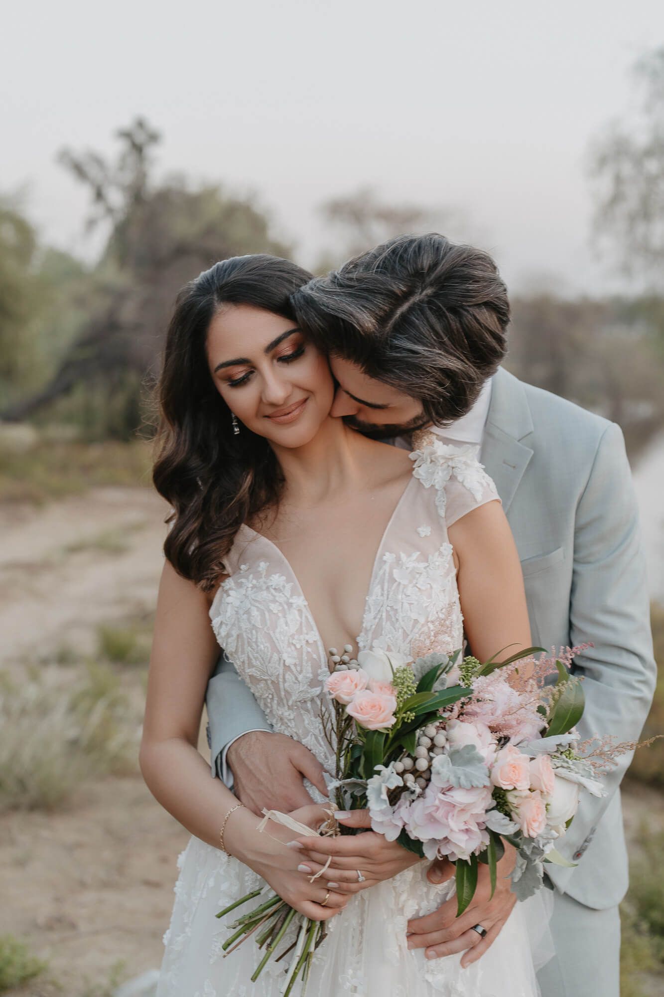 A couple shares an intimate moment in dusty outdoor setting wearing wedding attire and holding pink floral bouquet.