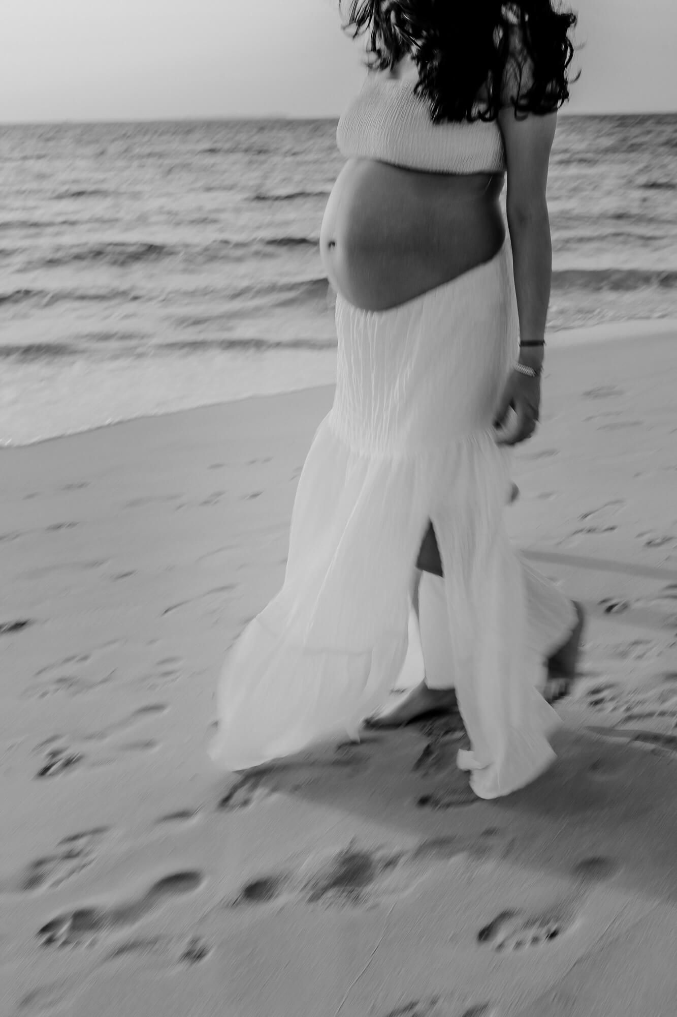 Black and white photo of a pregnant figure in a flowing white dress walking on a beach by the ocean.