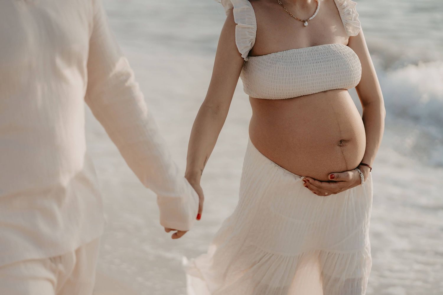 Intimate moment captured of an expecting couple in white attire holding hands on a beach.